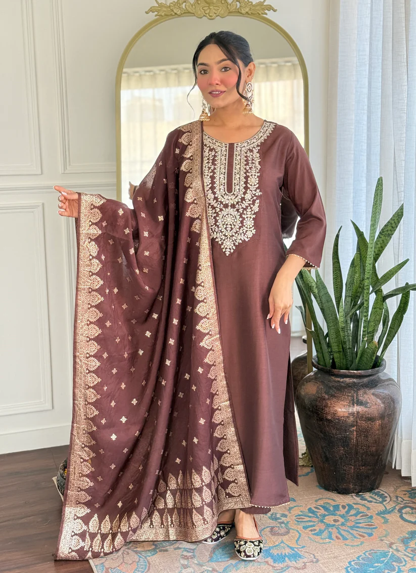 Woman in a brown dress with intricate patterns standing in a room with a mirror and plant.
