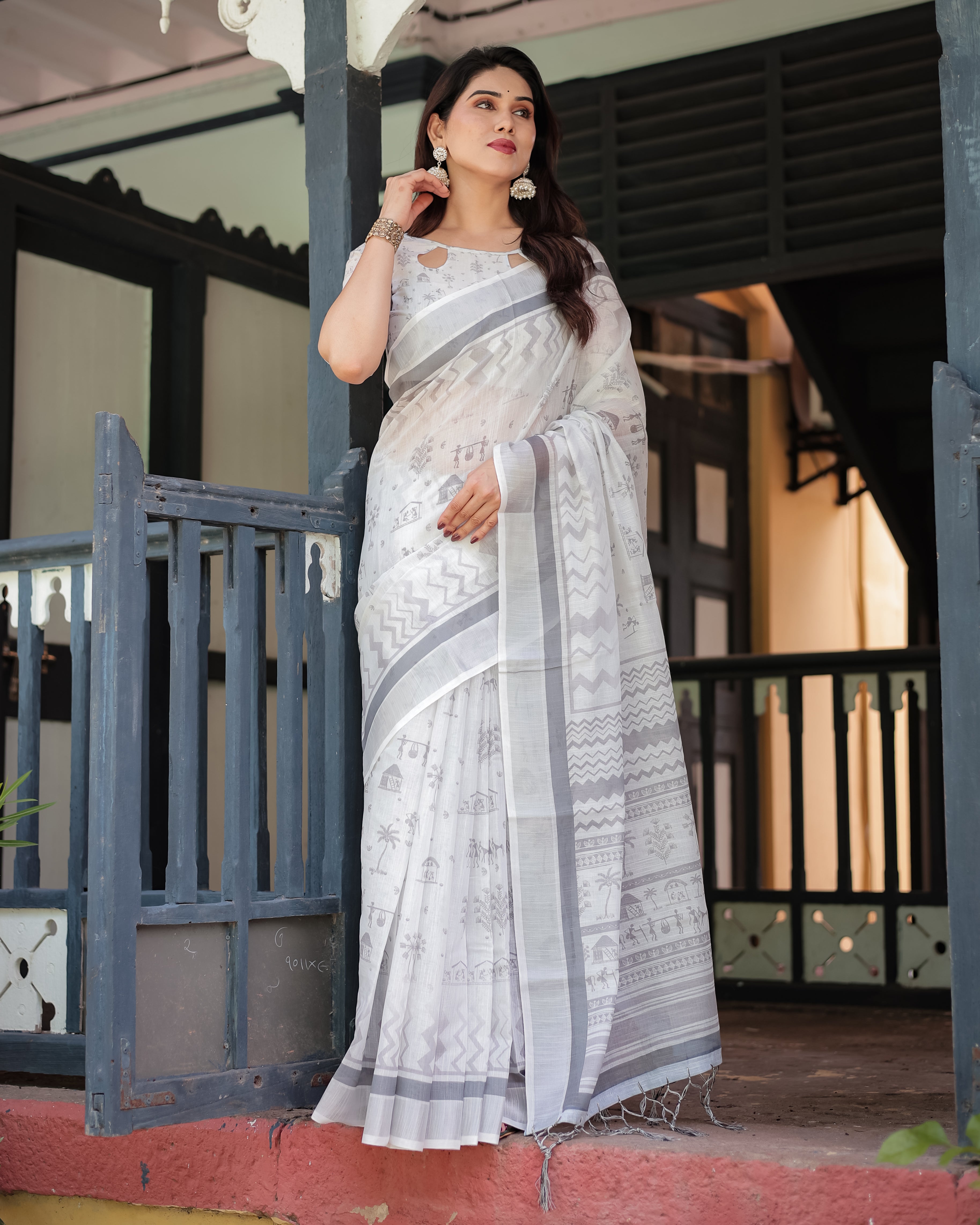 Woman in a white saree with gray patterns standing in front of a traditional building.