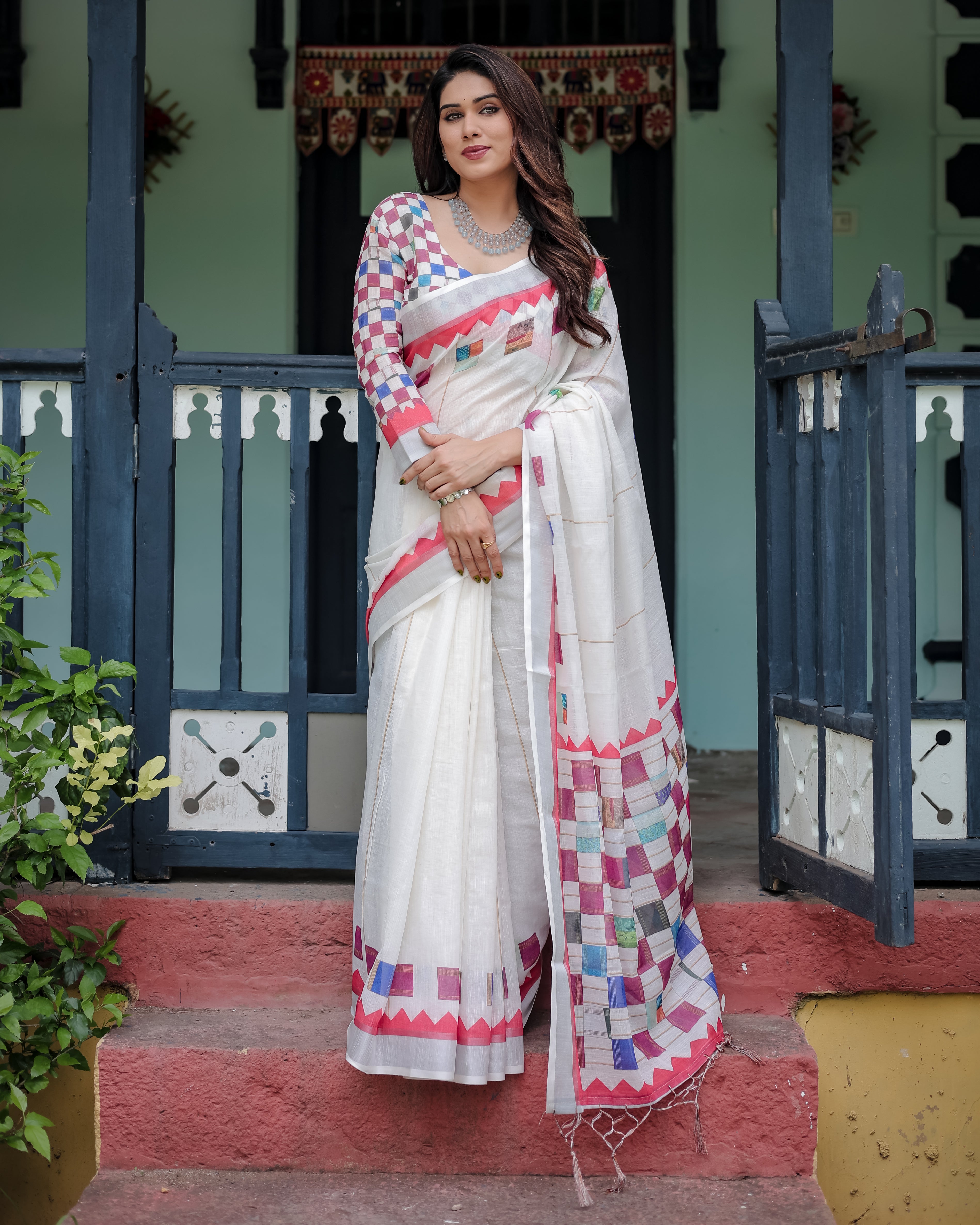 Woman in a white saree with colorful patterns standing in front of a traditional house.