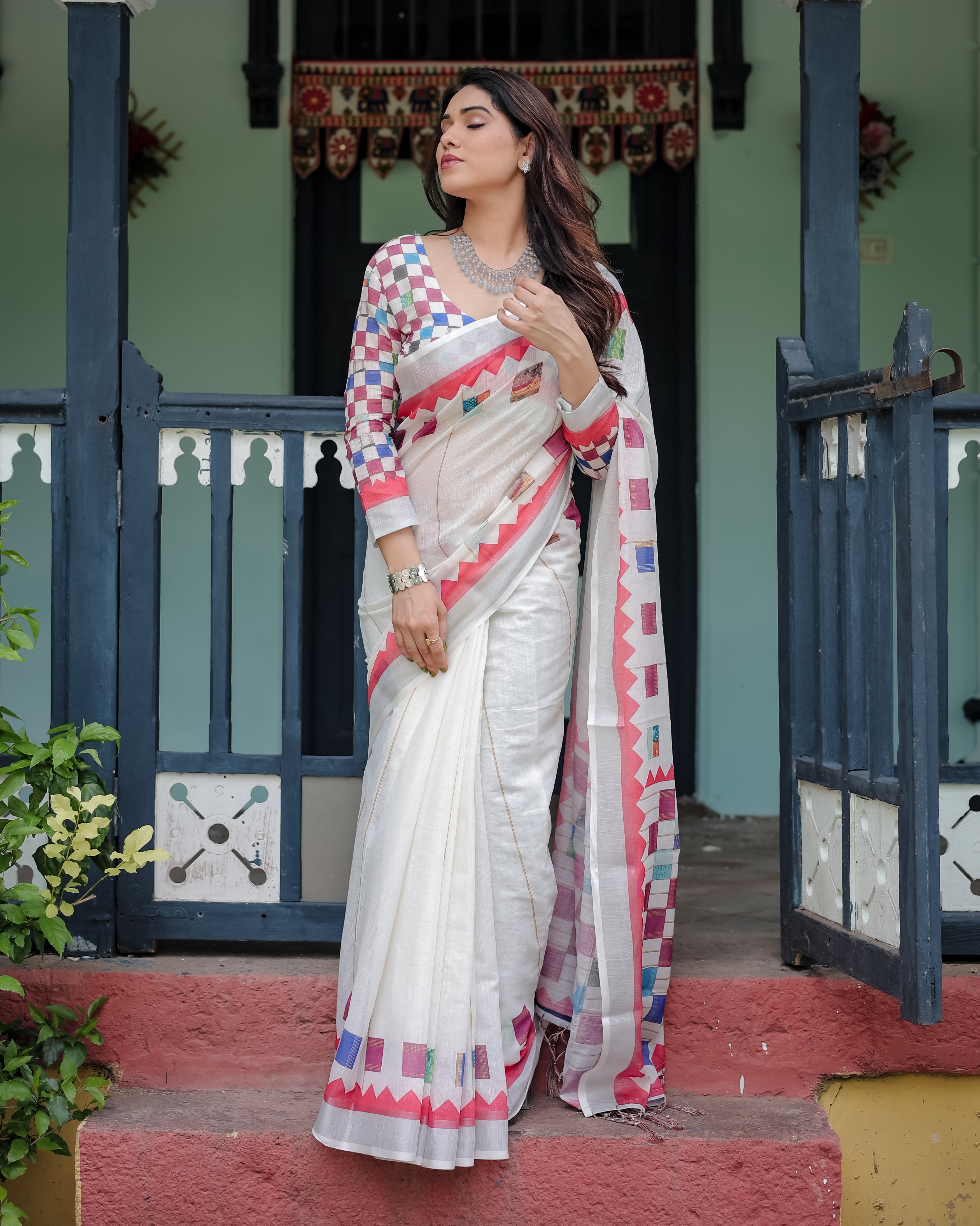 Woman in a white saree with colorful patterns standing in front of a traditional building.