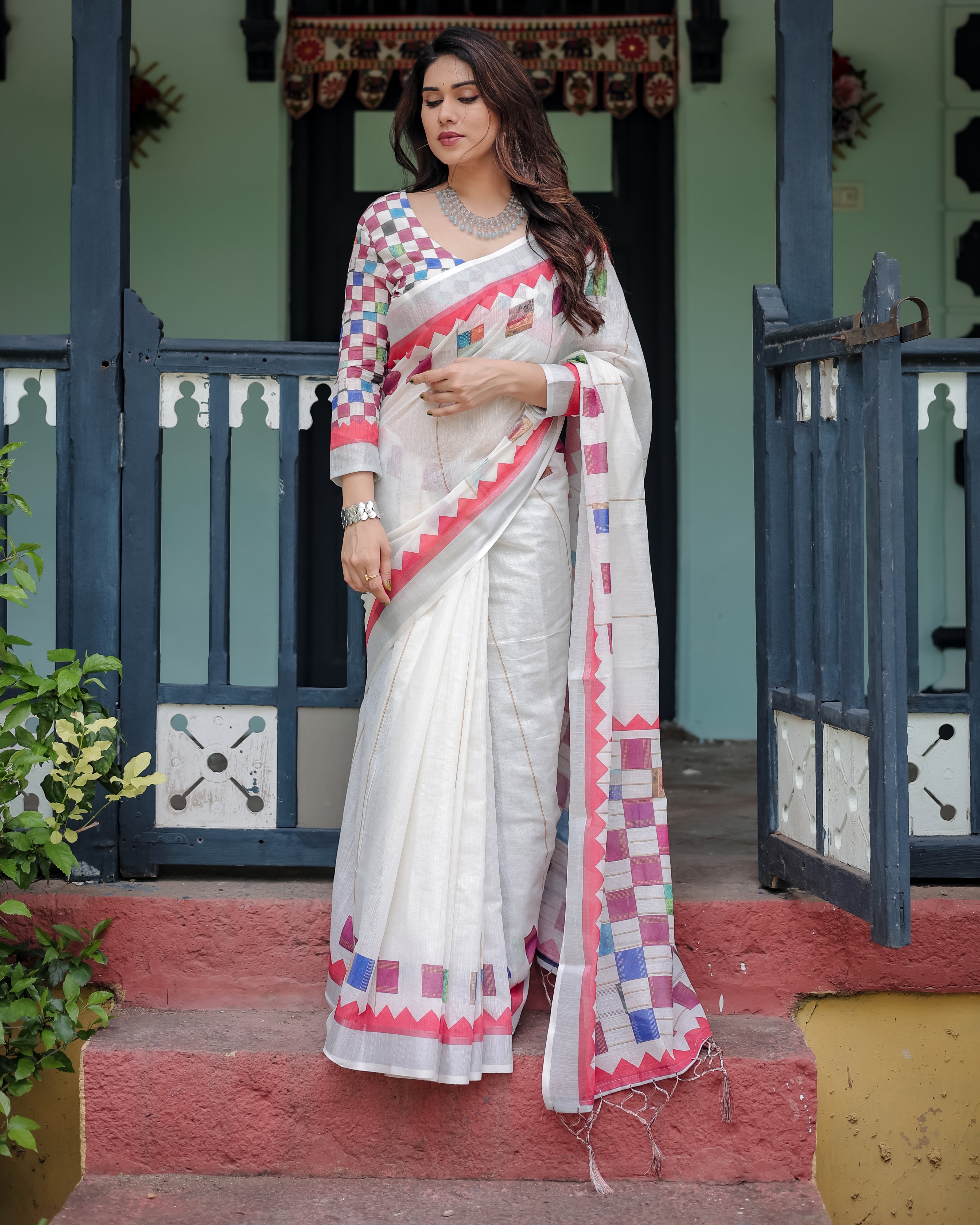 Woman wearing a white saree with colorful patterns standing in front of a traditional building.