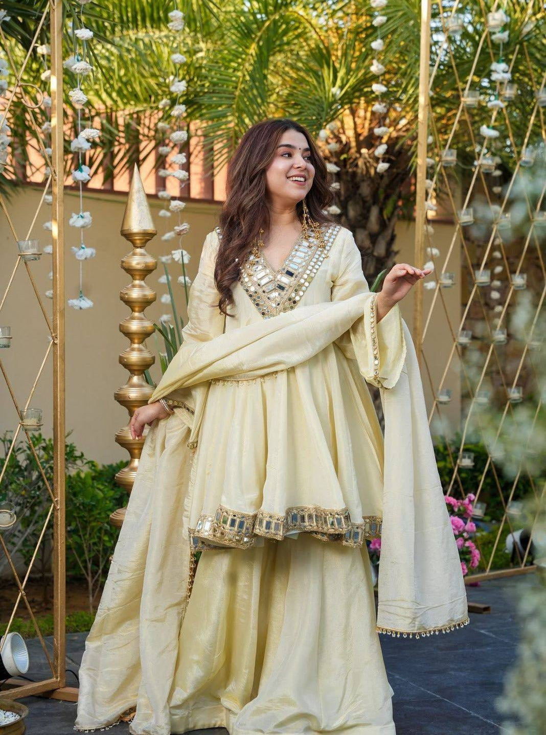Woman in a cream traditional Kurta Plazo Set outfit standing outdoors with decorative elements.