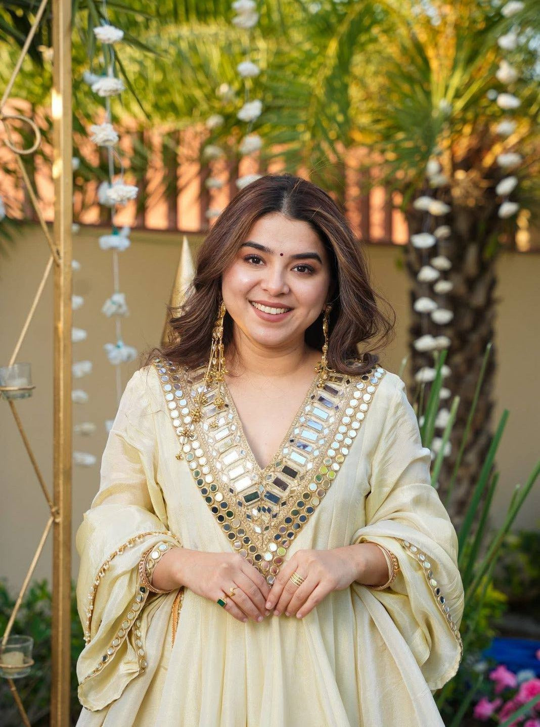 Woman in a traditional Kurta Plazo Set outfit with decorative elements, standing outdoors with plants and decorations in the background.