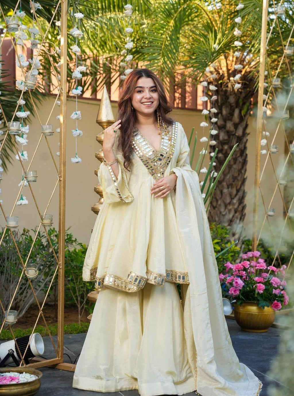 Woman in a cream traditional Kurta Plazo Set outfit with gold details standing outdoors with decorative elements.