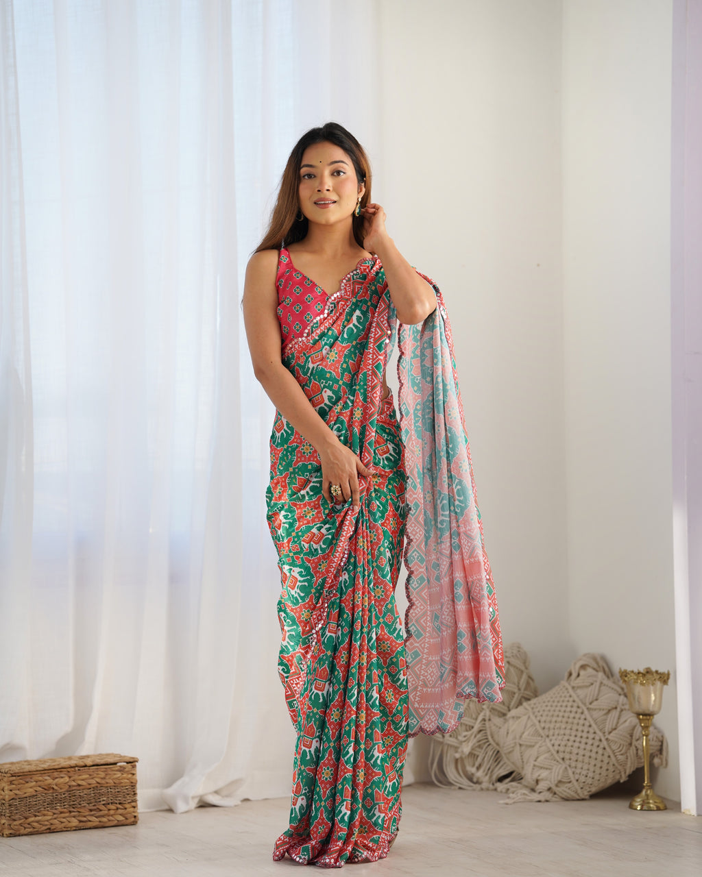 Woman wearing a floral saree standing in a room with white curtains and a woven basket.