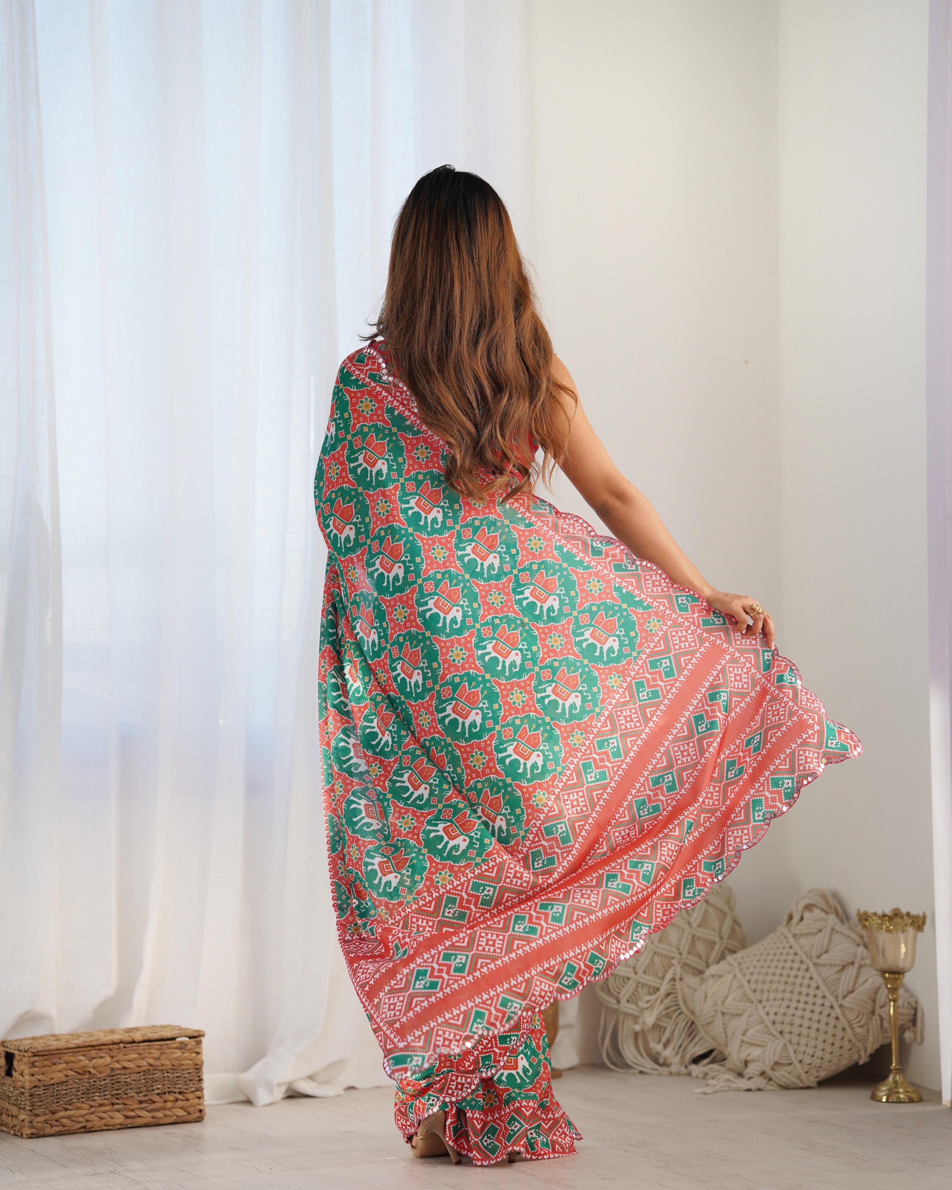 Woman wearing a colorful patterned saree in a bright room with white curtains.