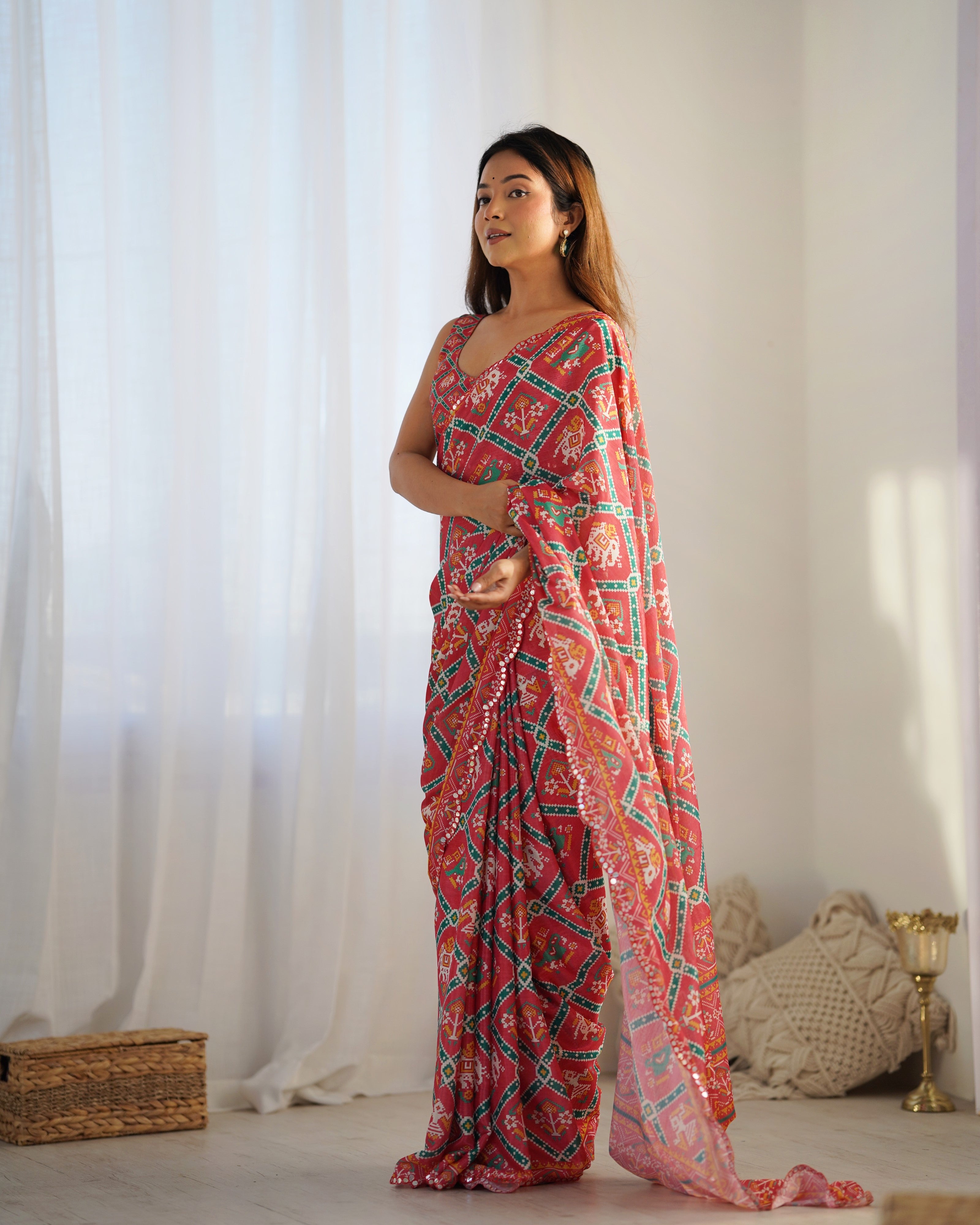 Woman wearing a pink patterned saree standing in a room with white curtains and a textured pillow.