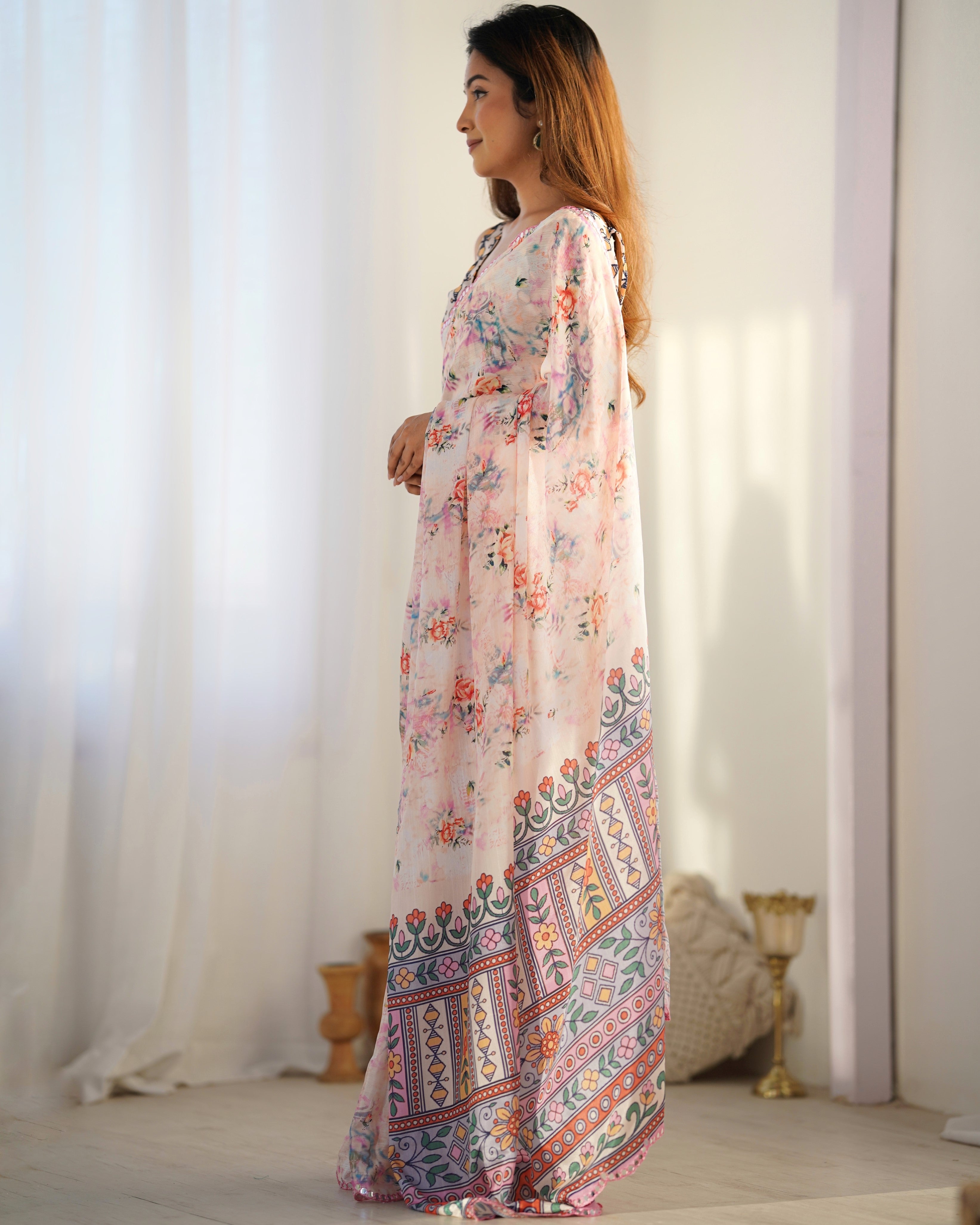 Woman wearing a long, floral-patterned saree with a colorful border against a white curtain background.