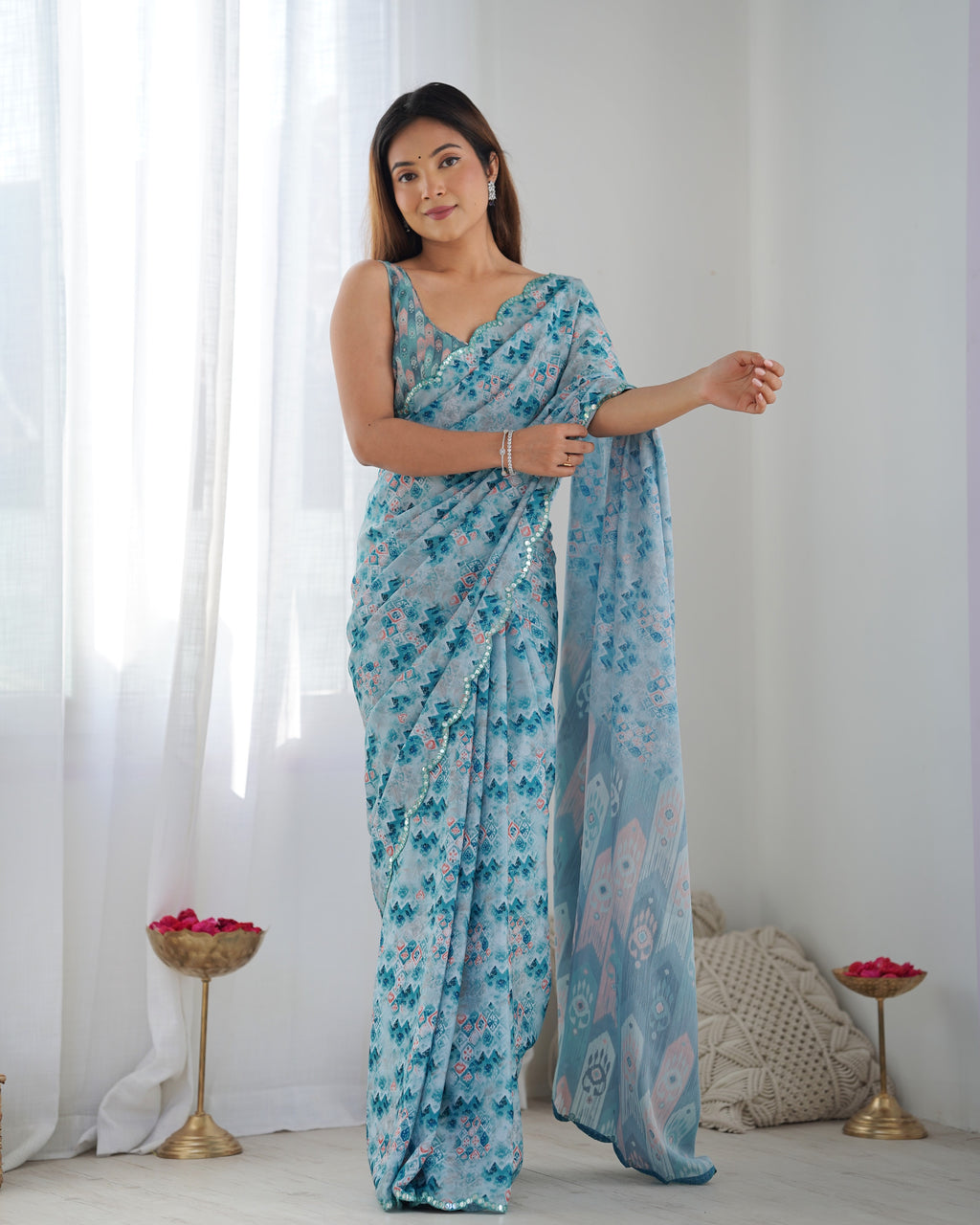 Woman wearing a blue floral saree in a bright room with white curtains.