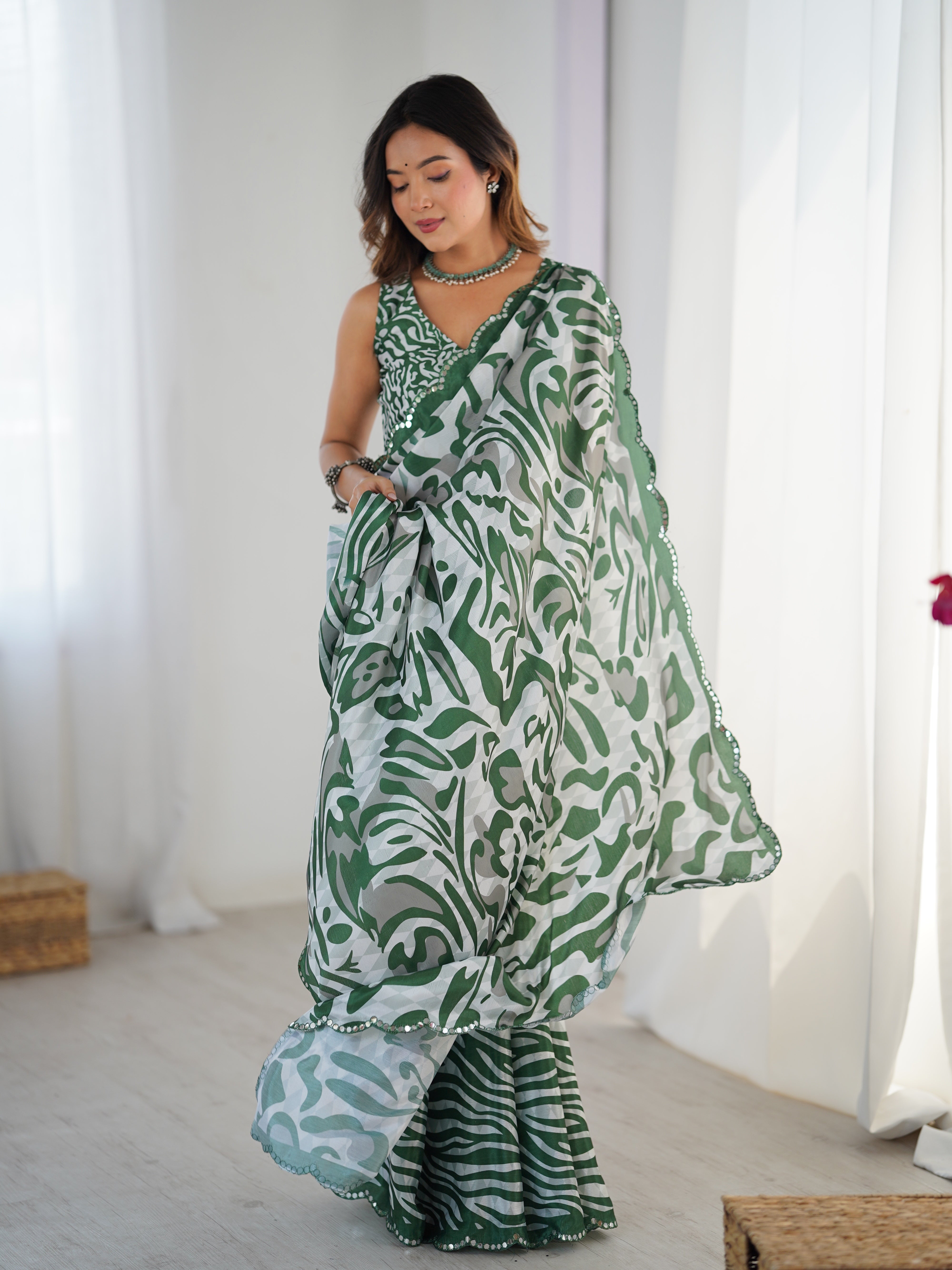 Woman wearing a green and white patterned saree in a bright room with white curtains.