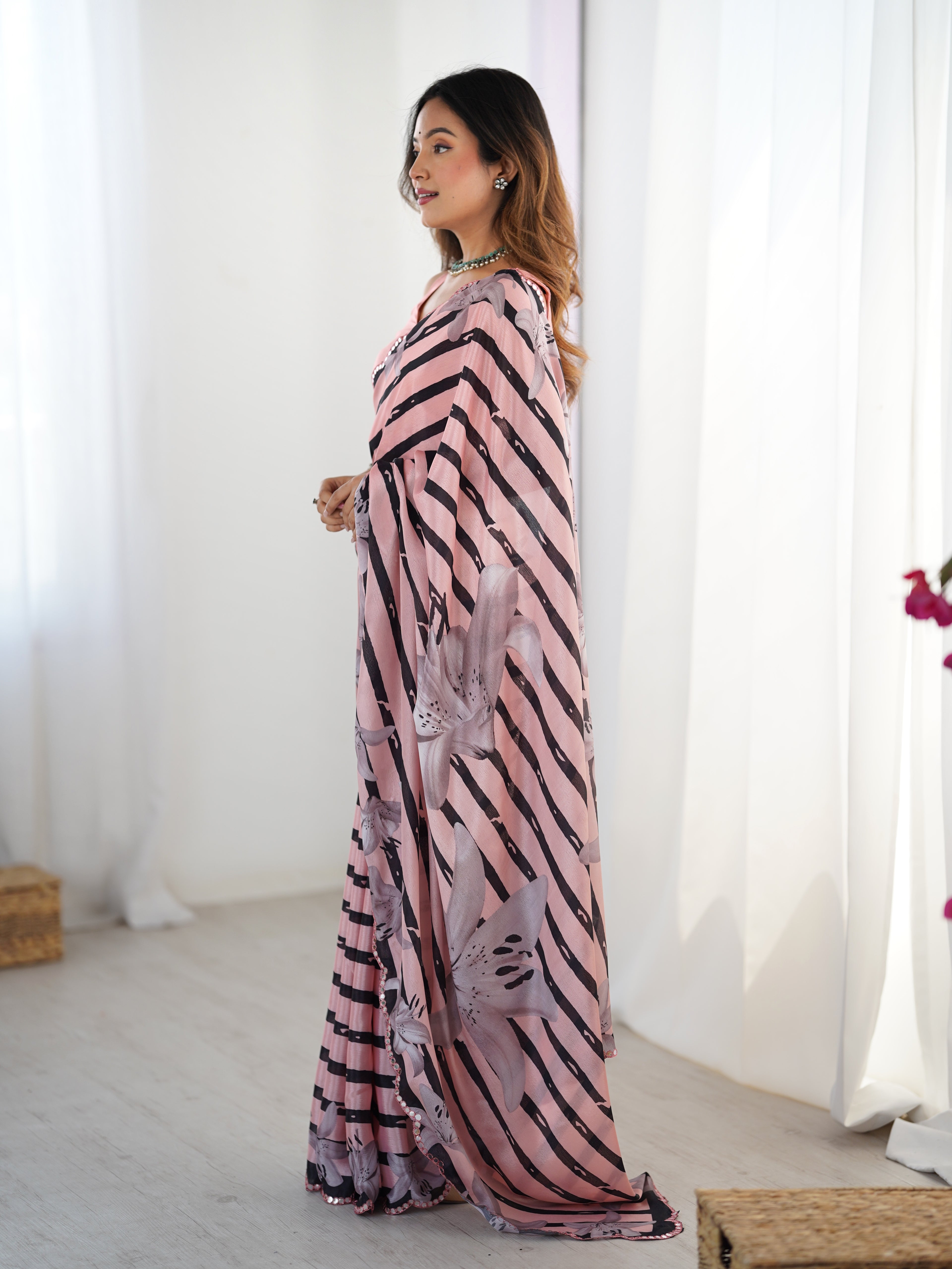 Woman wearing a long, patterned saree standing in a bright room with white curtains.