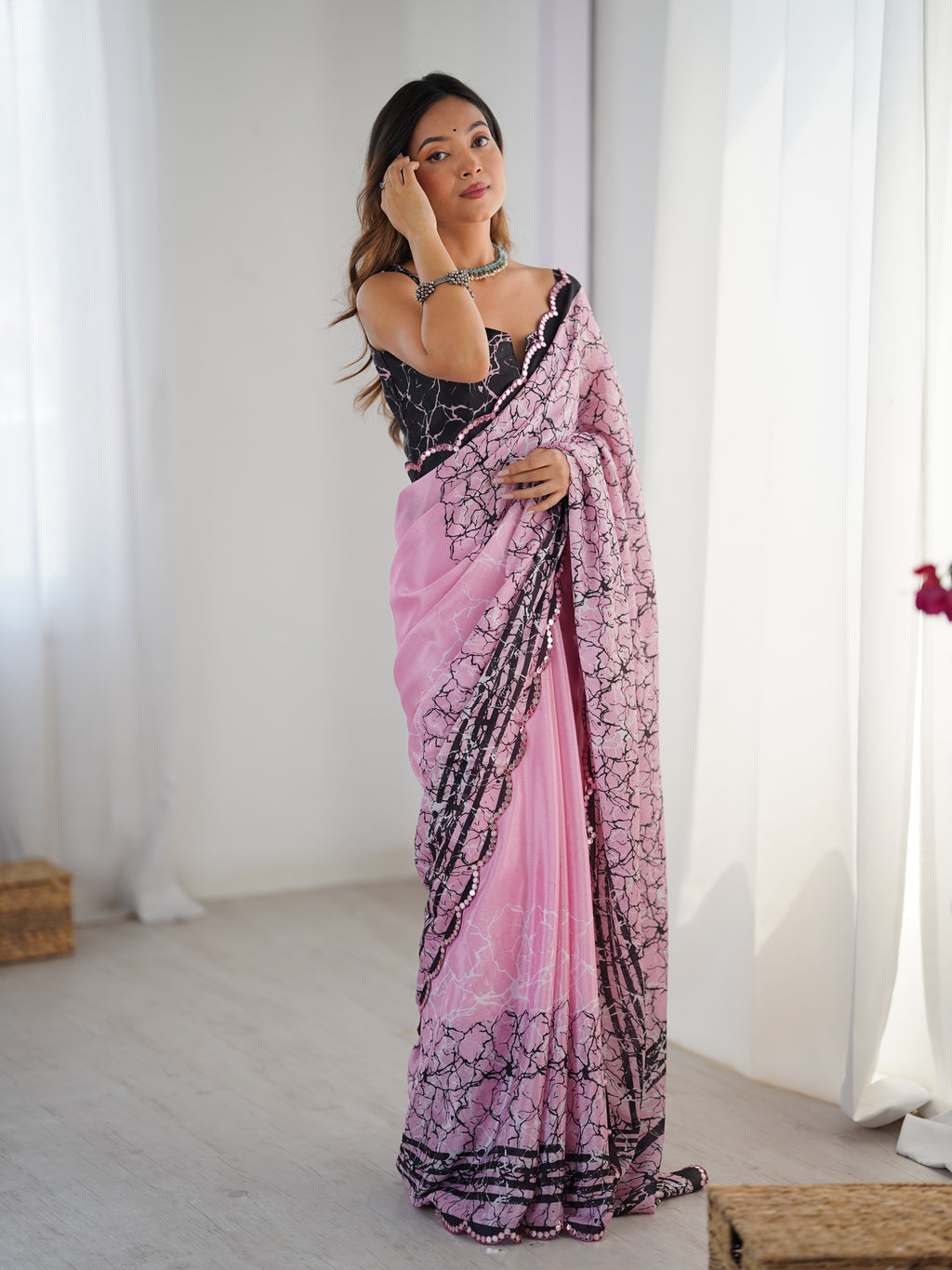 Woman wearing a pink and black saree in a bright room with white curtains.
