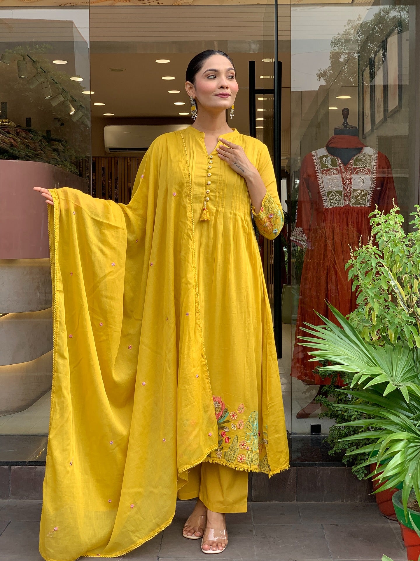 Woman in a yellow kurta set outfit standing in front of a store with a plant in the foreground.