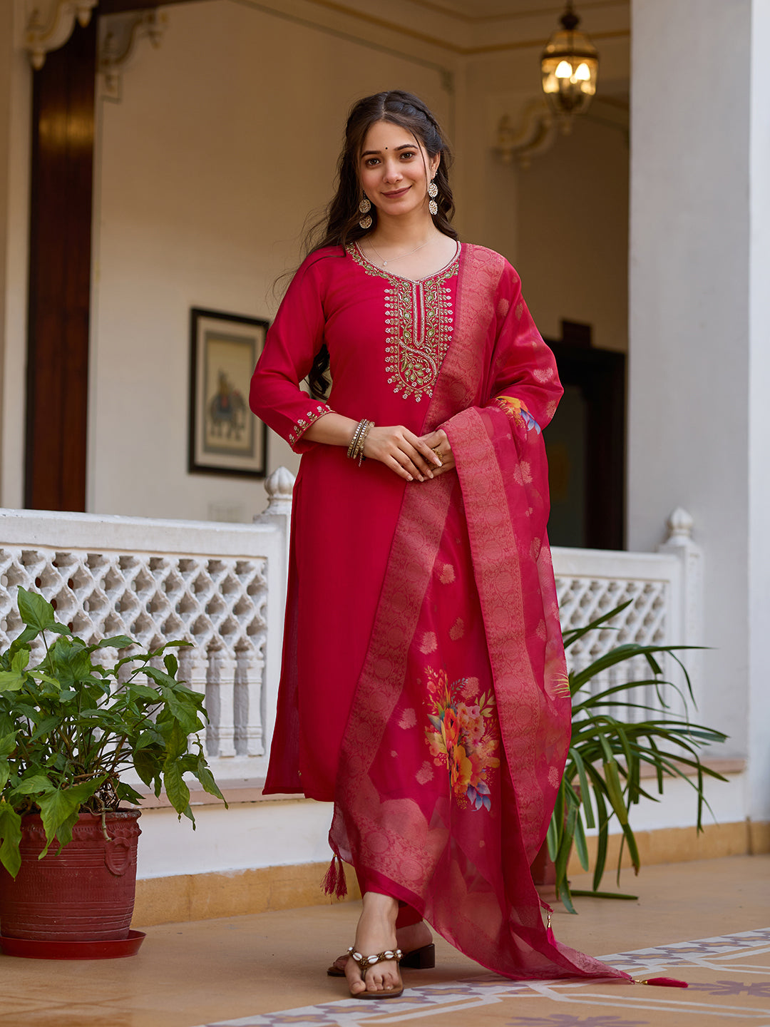 Woman in a red traditional outfit standing indoors with plants and a decorative wall in the background