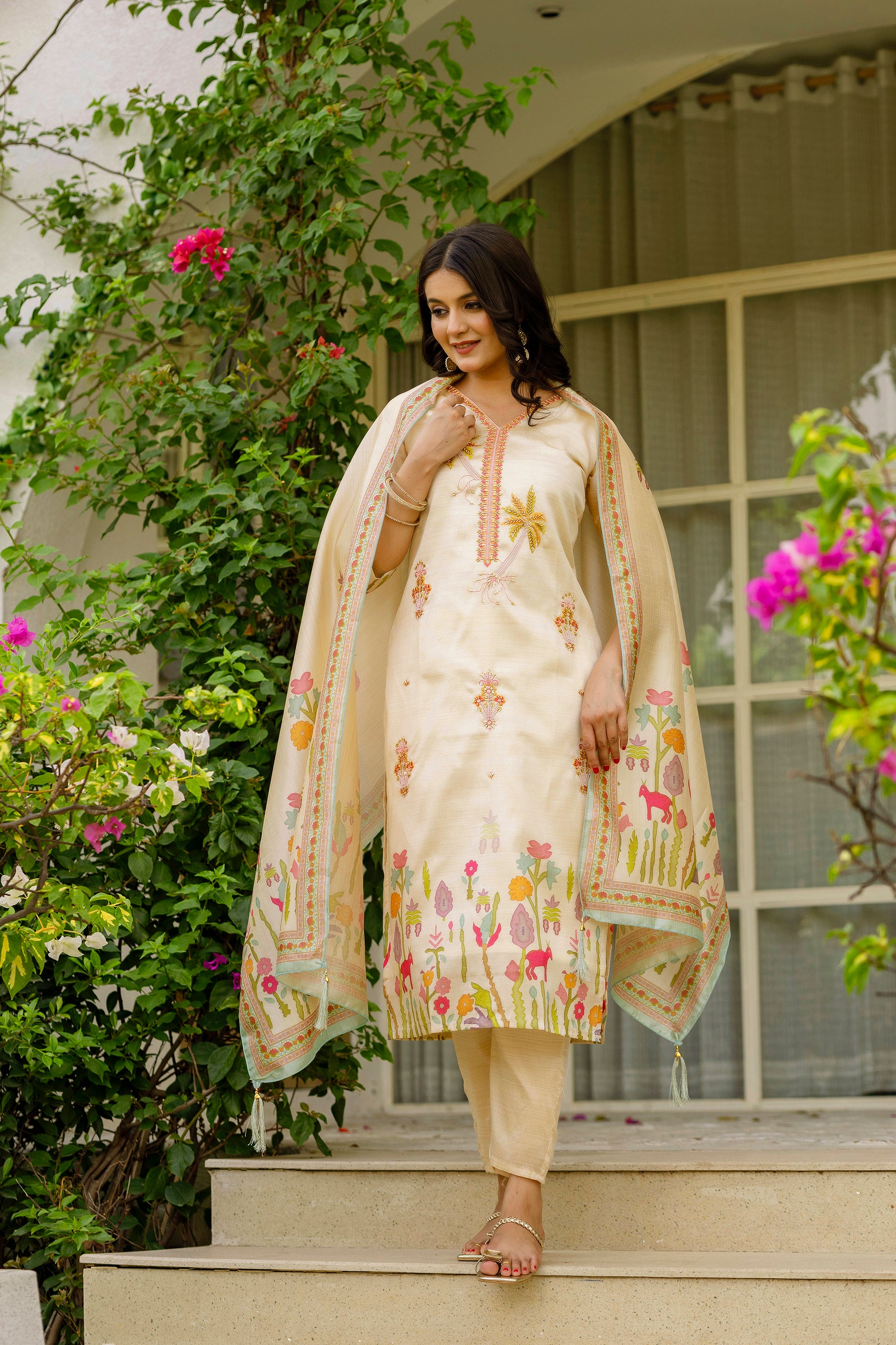 Woman in a cream-colored traditional outfit with floral patterns standing on steps outdoors.