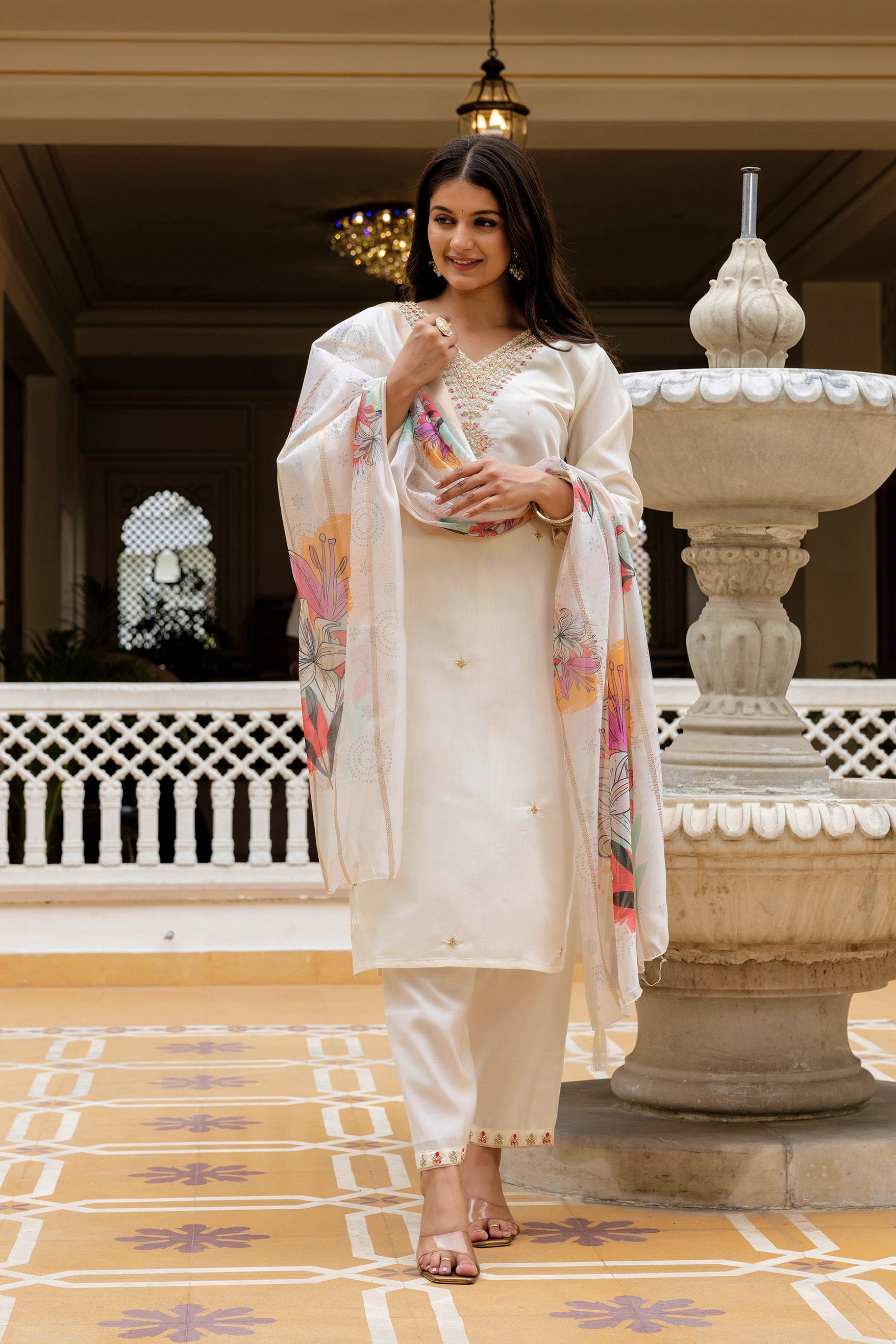 Woman in a white traditional outfit with a colorful scarf standing in a decorative interior setting.