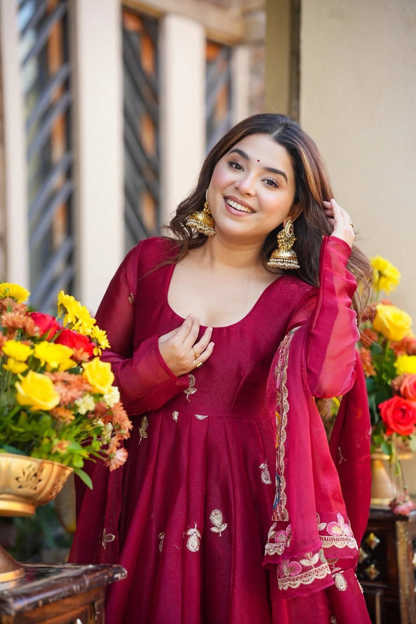 Woman in a red traditional Anarkali Palazzo Set outfit with floral decorations and jewelry, standing in front of a floral arrangement.
