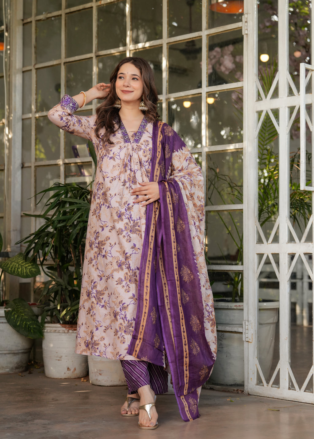 Woman in a floral dress with a purple dupatta standing in a decorative indoor setting.