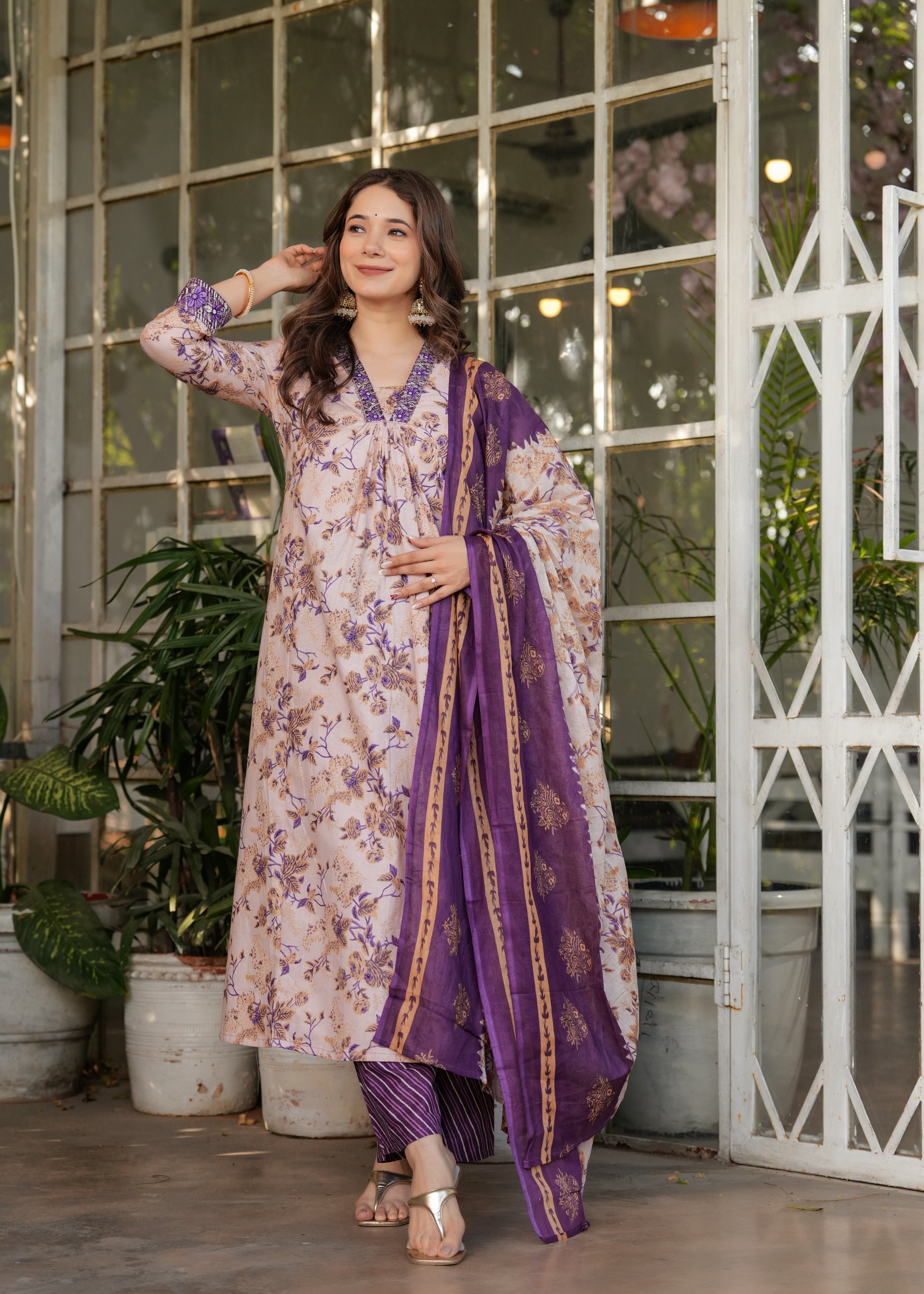 Woman in a floral dress with a purple dupatta standing in a decorative indoor setting.