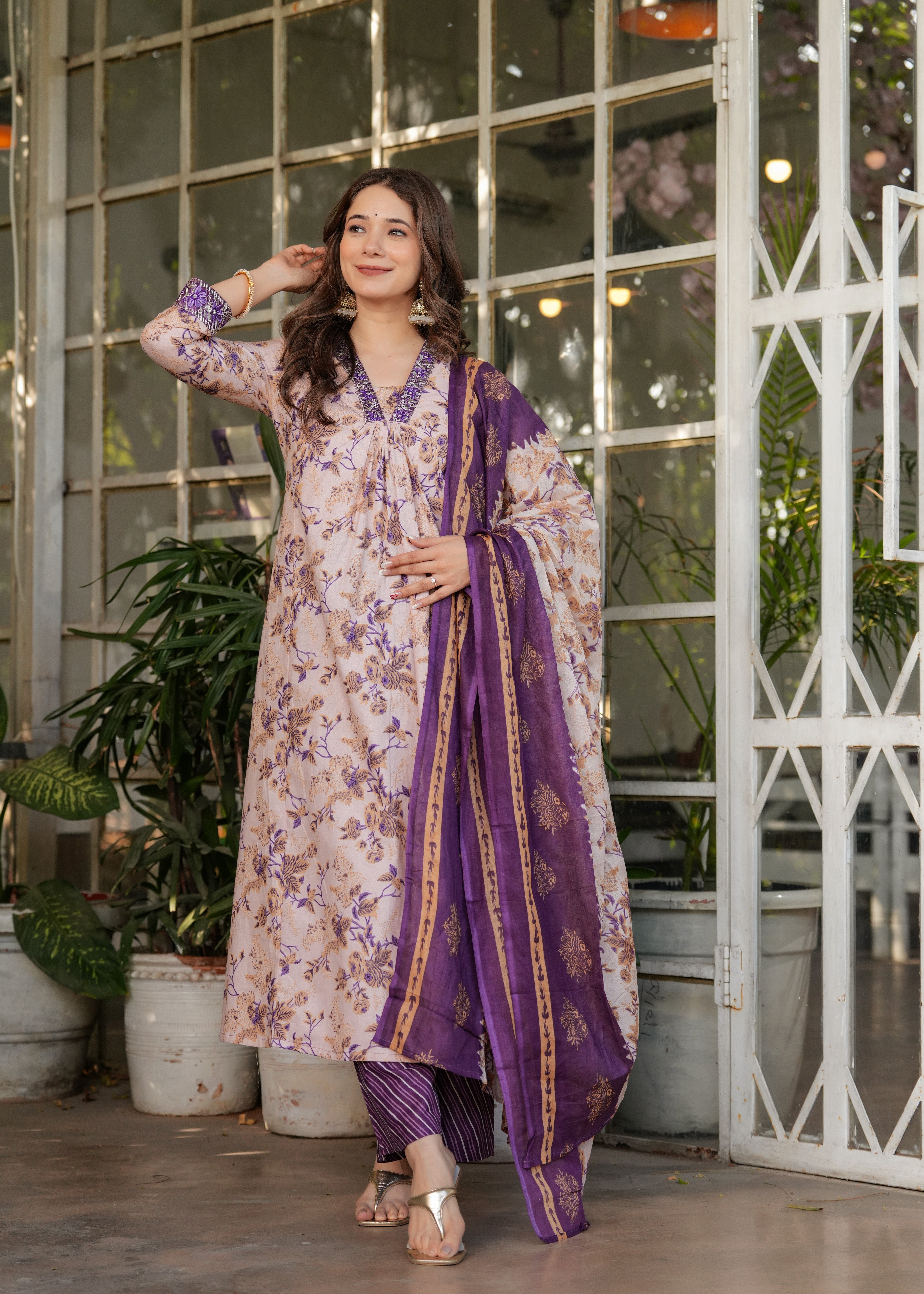 Woman in a floral dress with a purple dupatta standing in a decorative indoor setting.