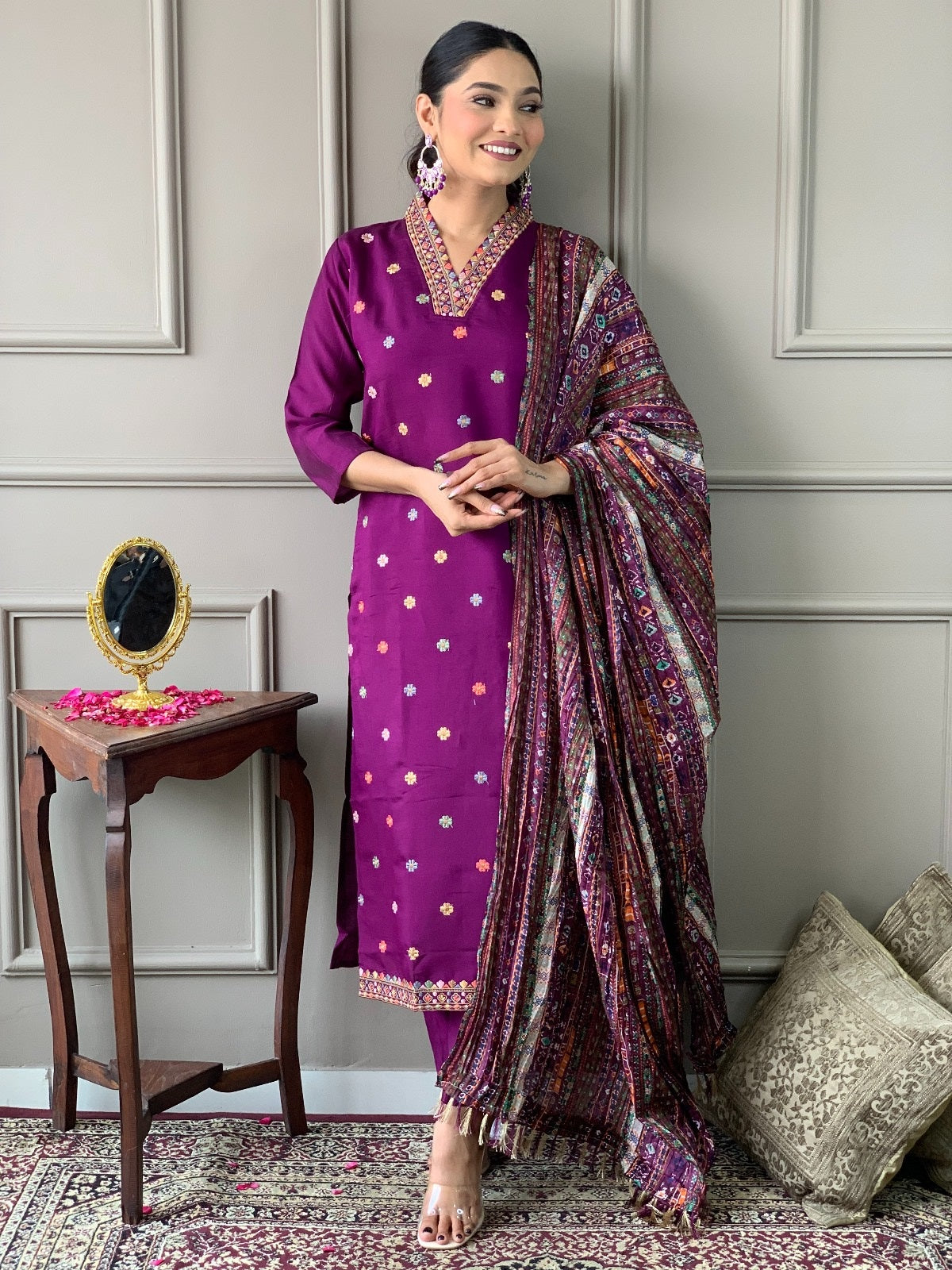 Woman in a purple traditional outfit with a patterned scarf standing in a room with decorative elements.