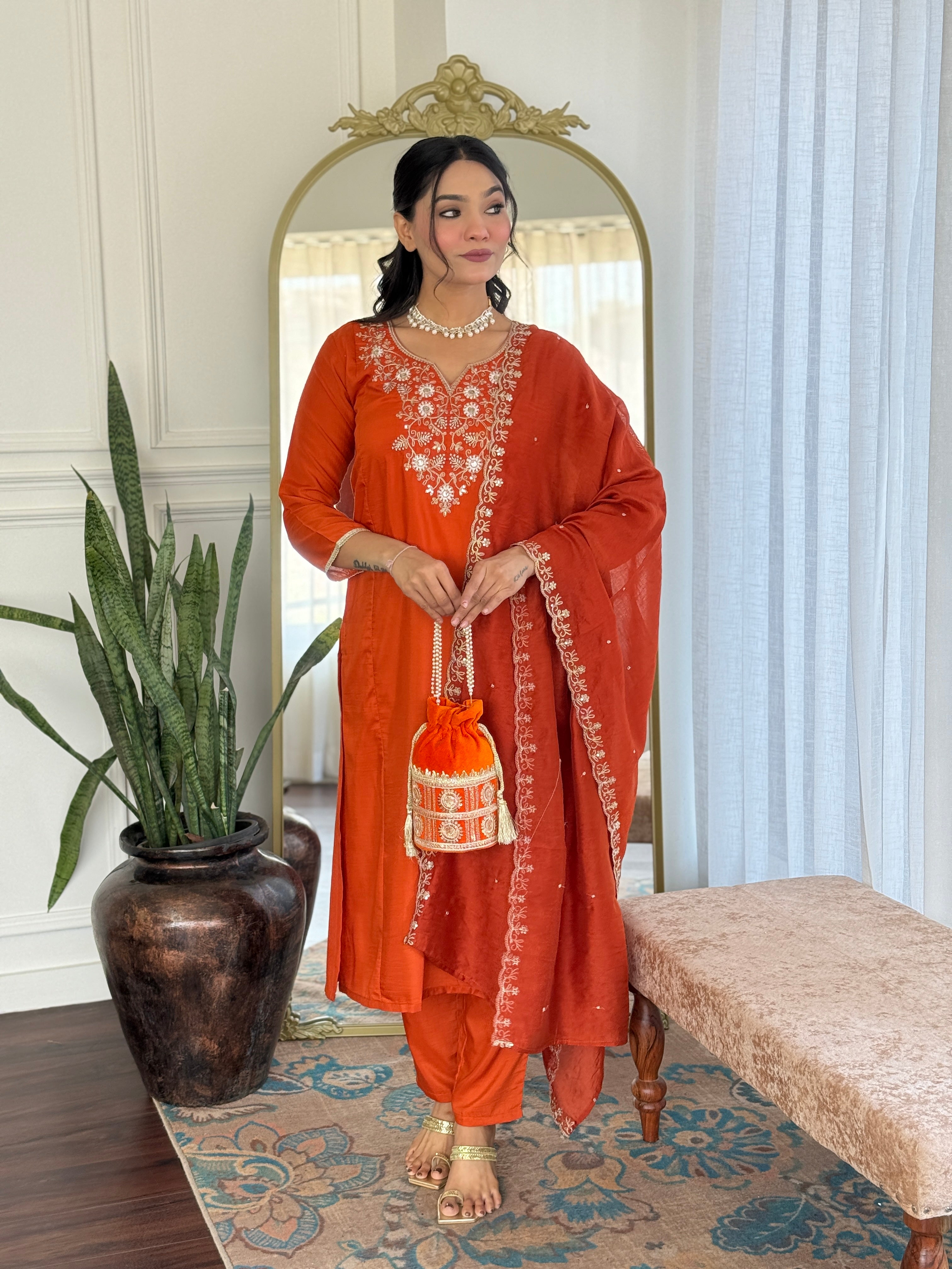 Woman in an orange traditional outfit standing in a room with a mirror and plant.