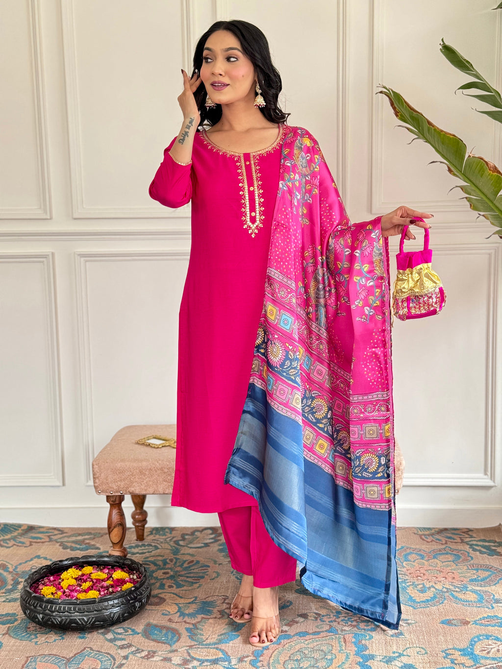 Woman in a pink traditional outfit with a matching dupatta, holding a small bag, against a white paneled wall.