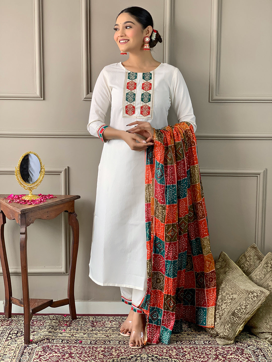 Woman in a white kurta with colorful patterned dupatta standing in a room with a mirror and pillows.