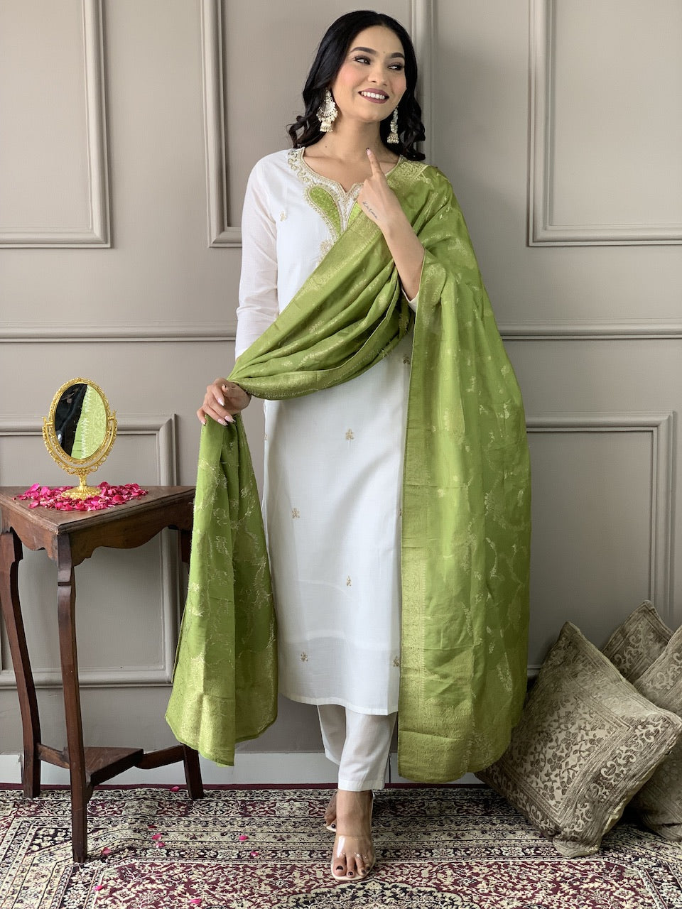 Woman in a white outfit with a green dupatta standing in a room with decorative elements.