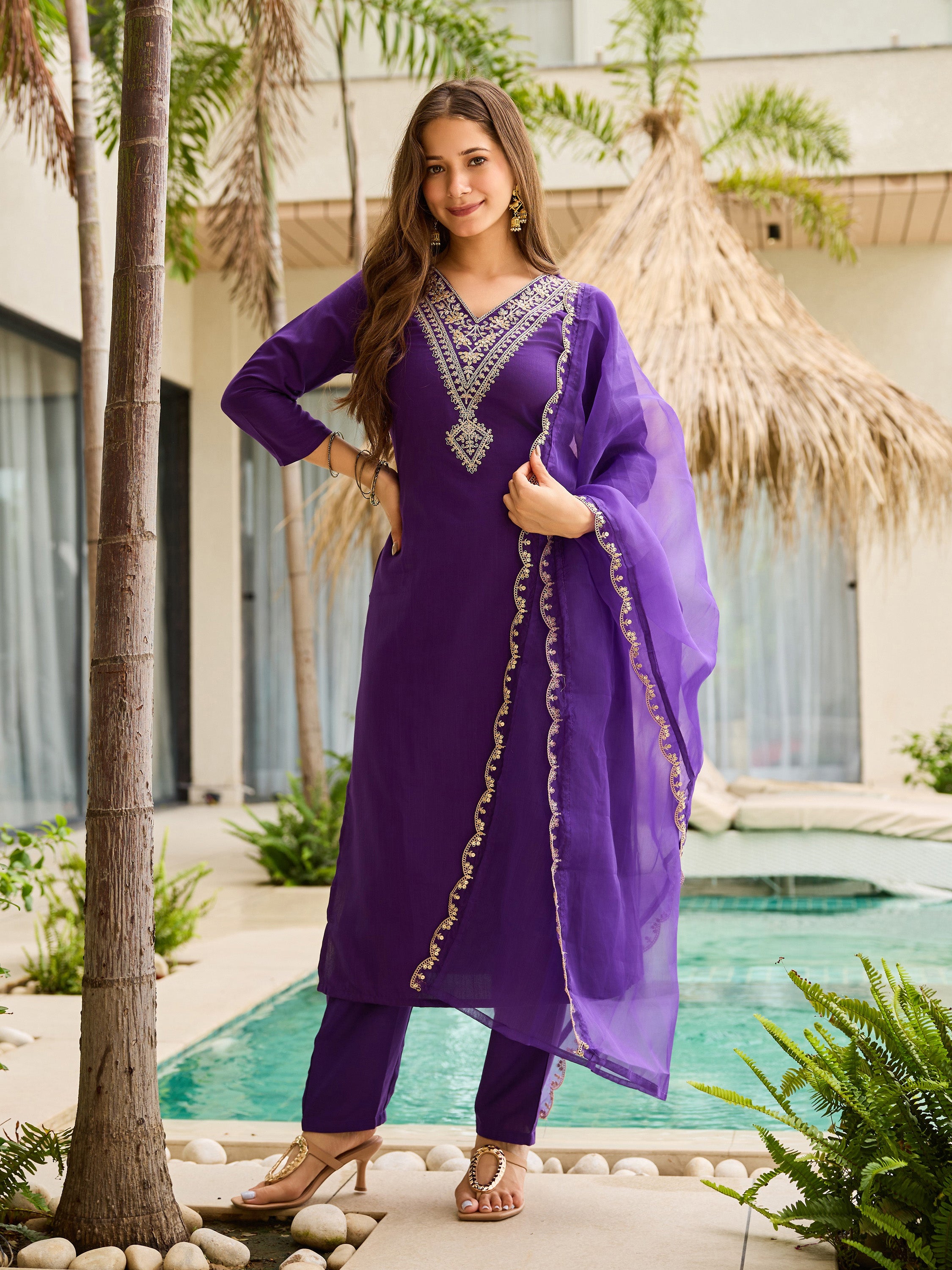 Woman in a purple kurta set traditional outfit standing by a pool with palm trees in the background