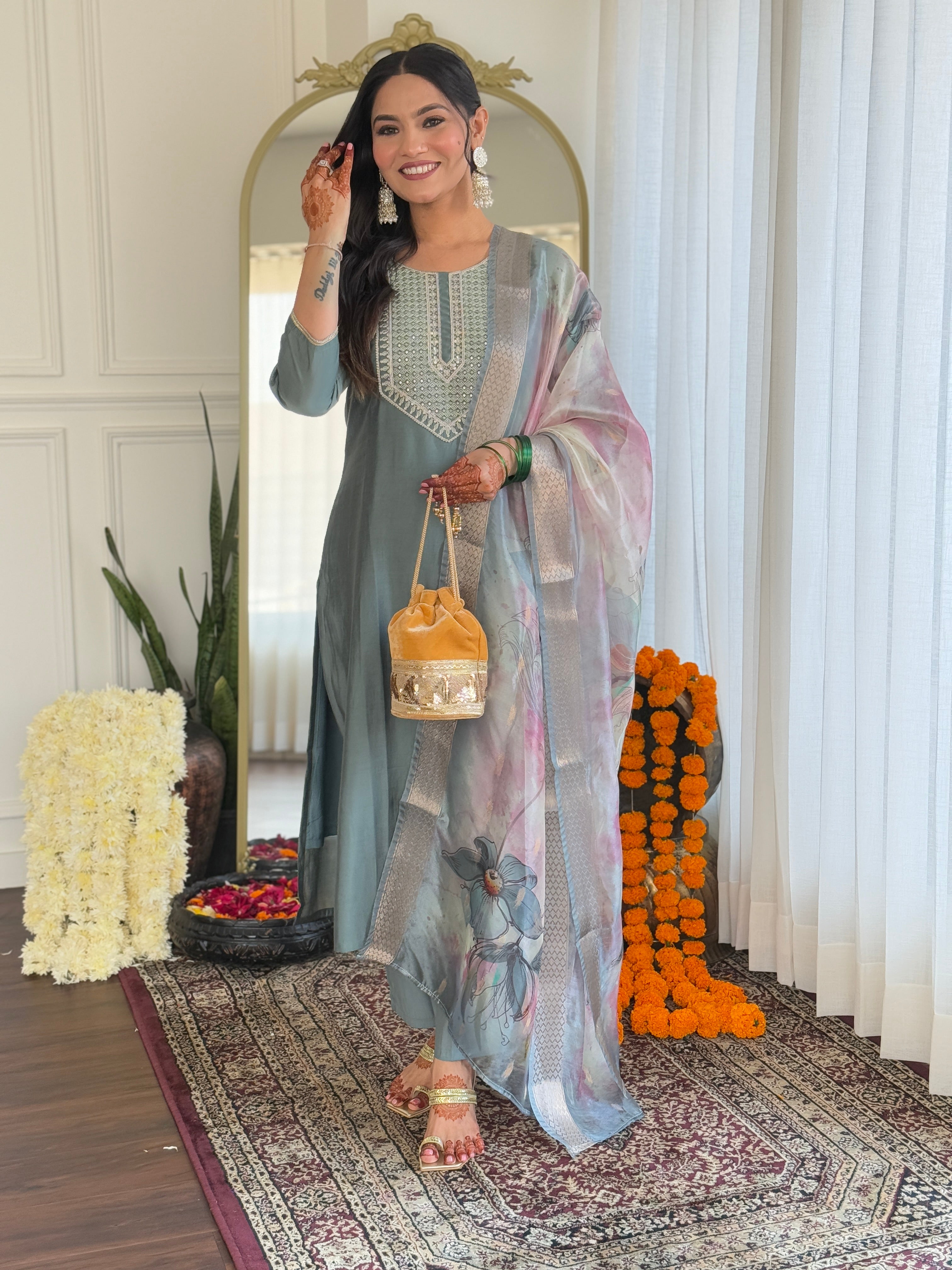 Woman in traditional outfit standing in a decorated room with flowers and a mirror.