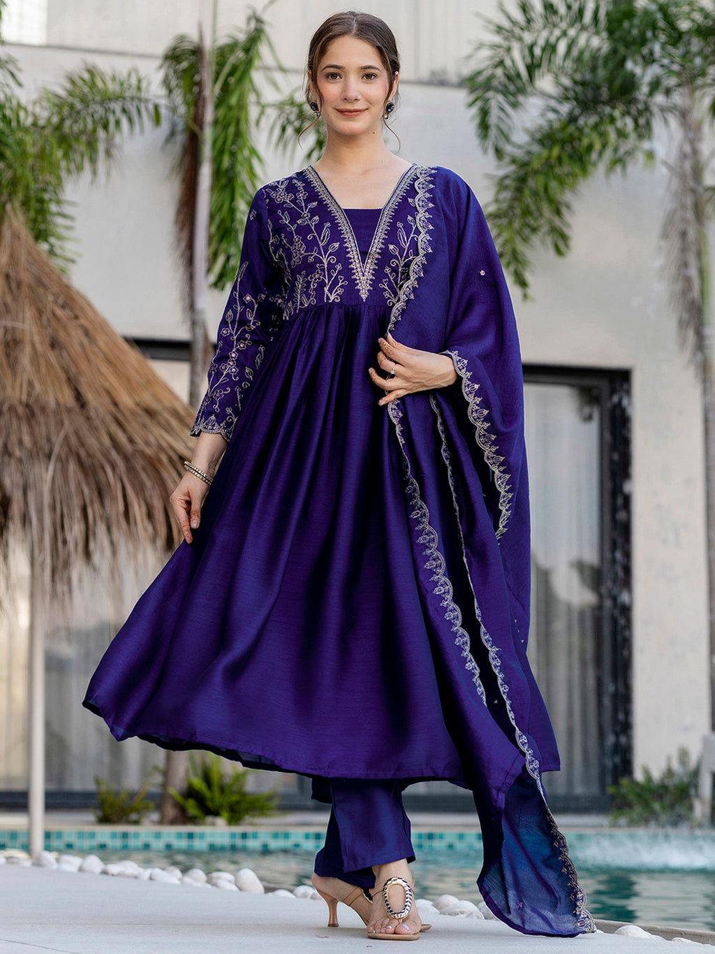 Woman wearing a purple kurta set with embroidered outfit standing by a pool with greenery in the background