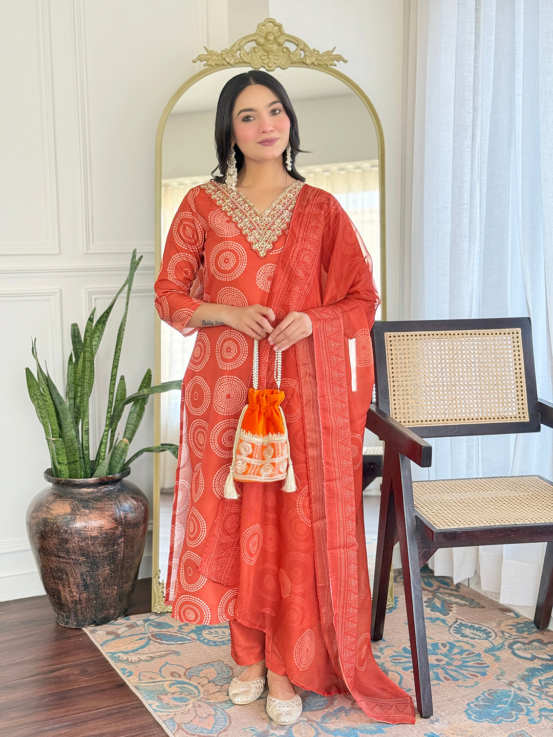 Woman in a red patterned outfit holding a matching bag in a room with a mirror and chair.