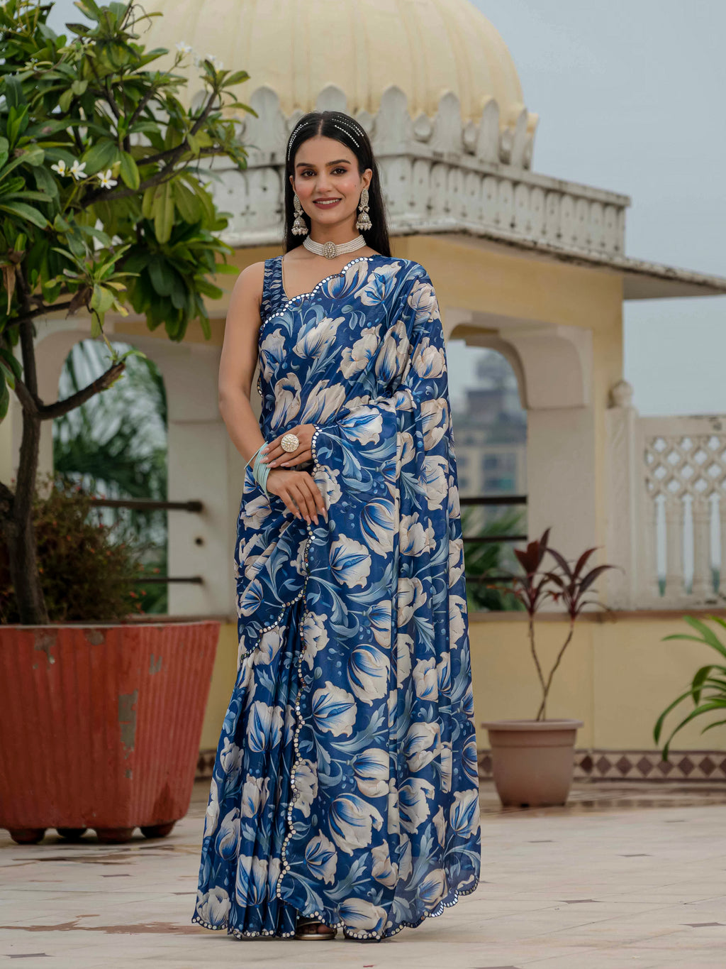 Woman in a blue ready to wear saree standing on a rooftop with plants and architectural elements in the background.