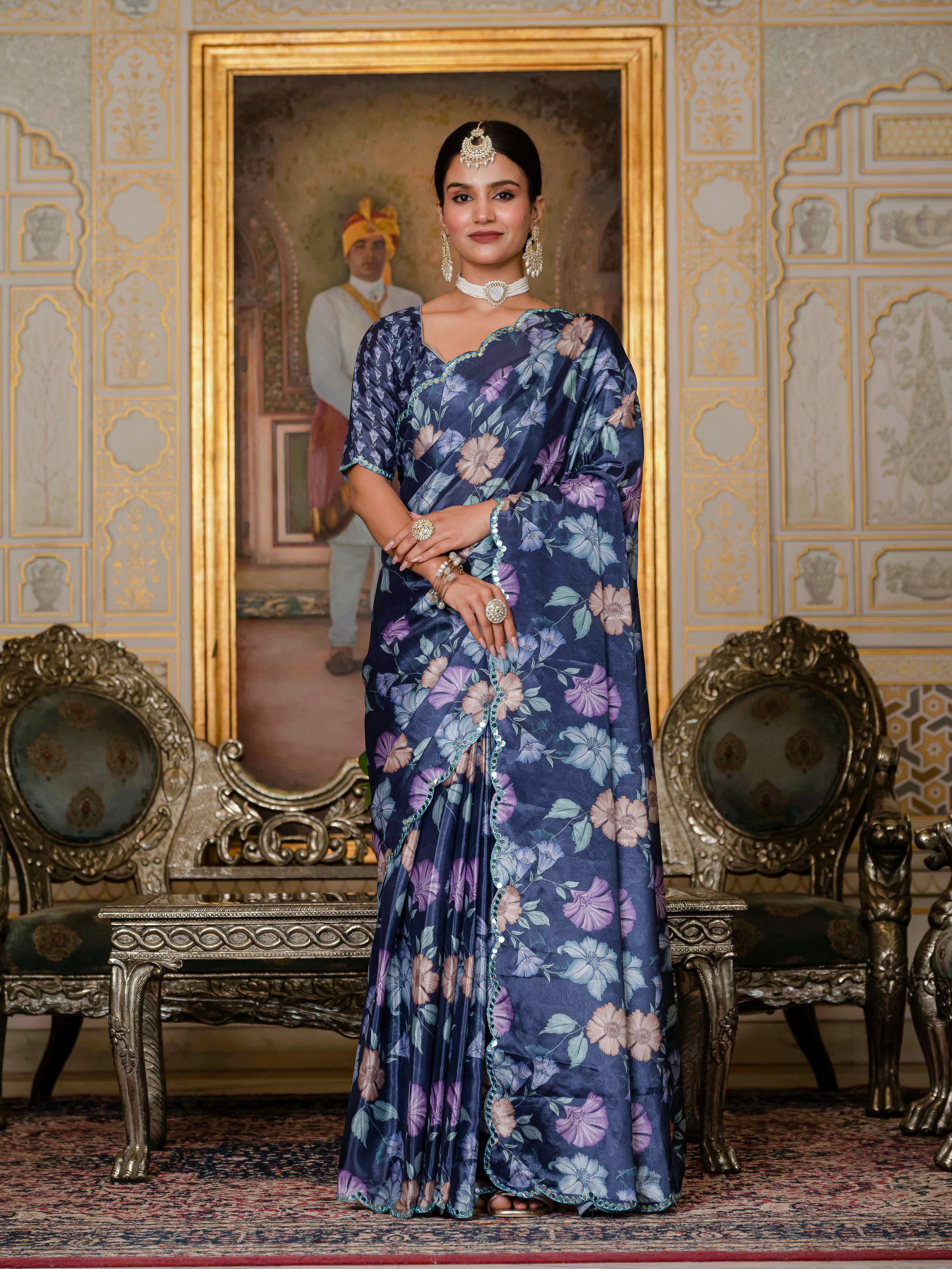 A woman wearing a navy blue floral ready to wear saree stands in an ornate room with decorative chairs and wall art.