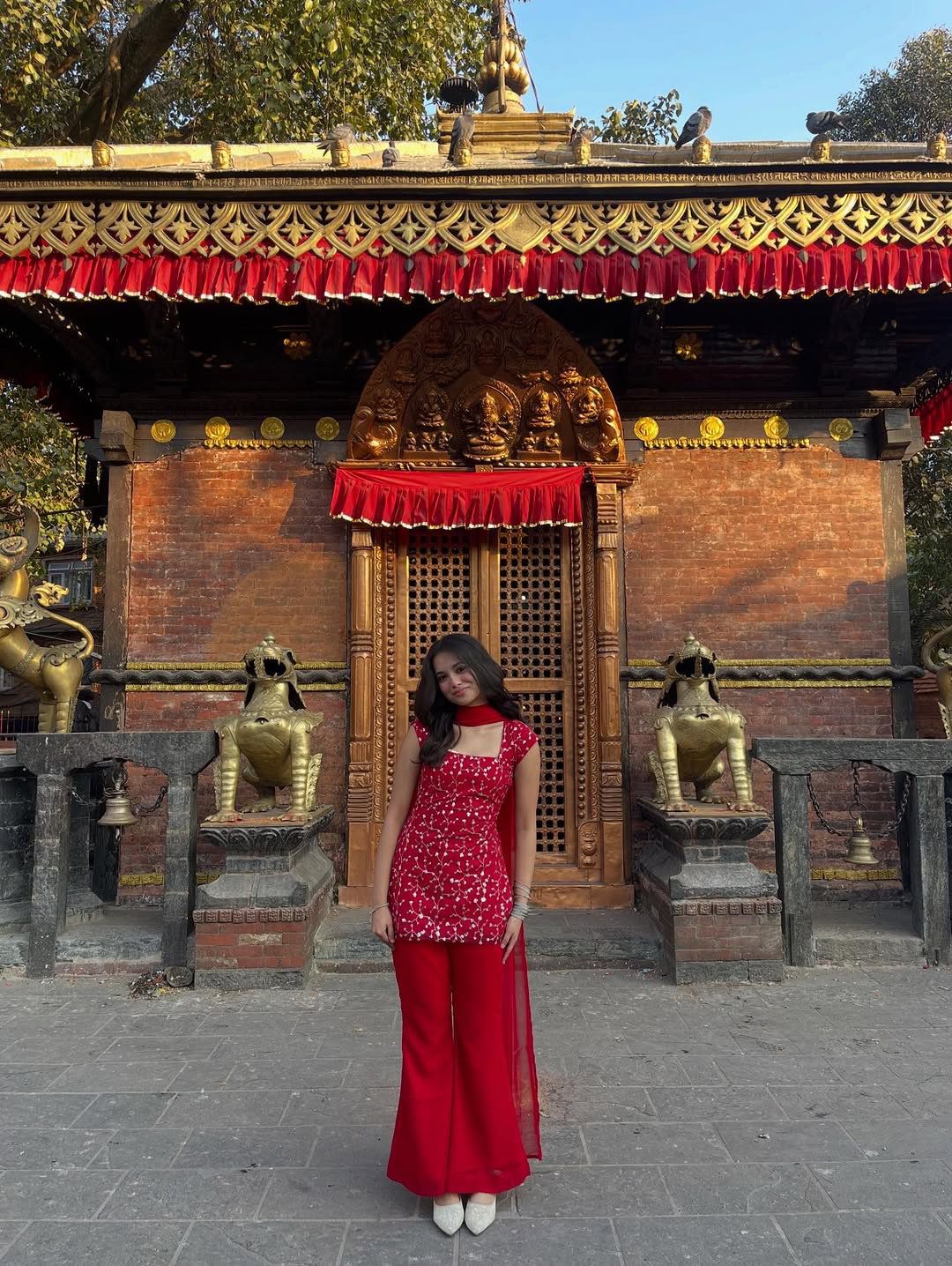 Woman in a red kurti standing in front of a temple with gold accents and red roofs.