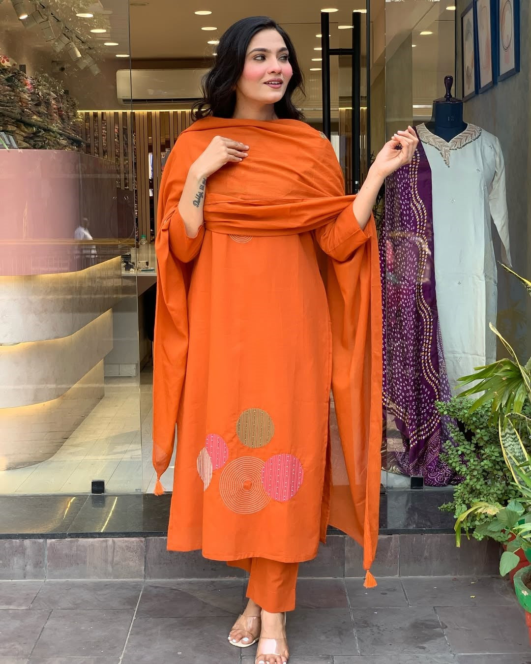 Woman in an orange kurta set outfit standing in a store with mannequins and plants.