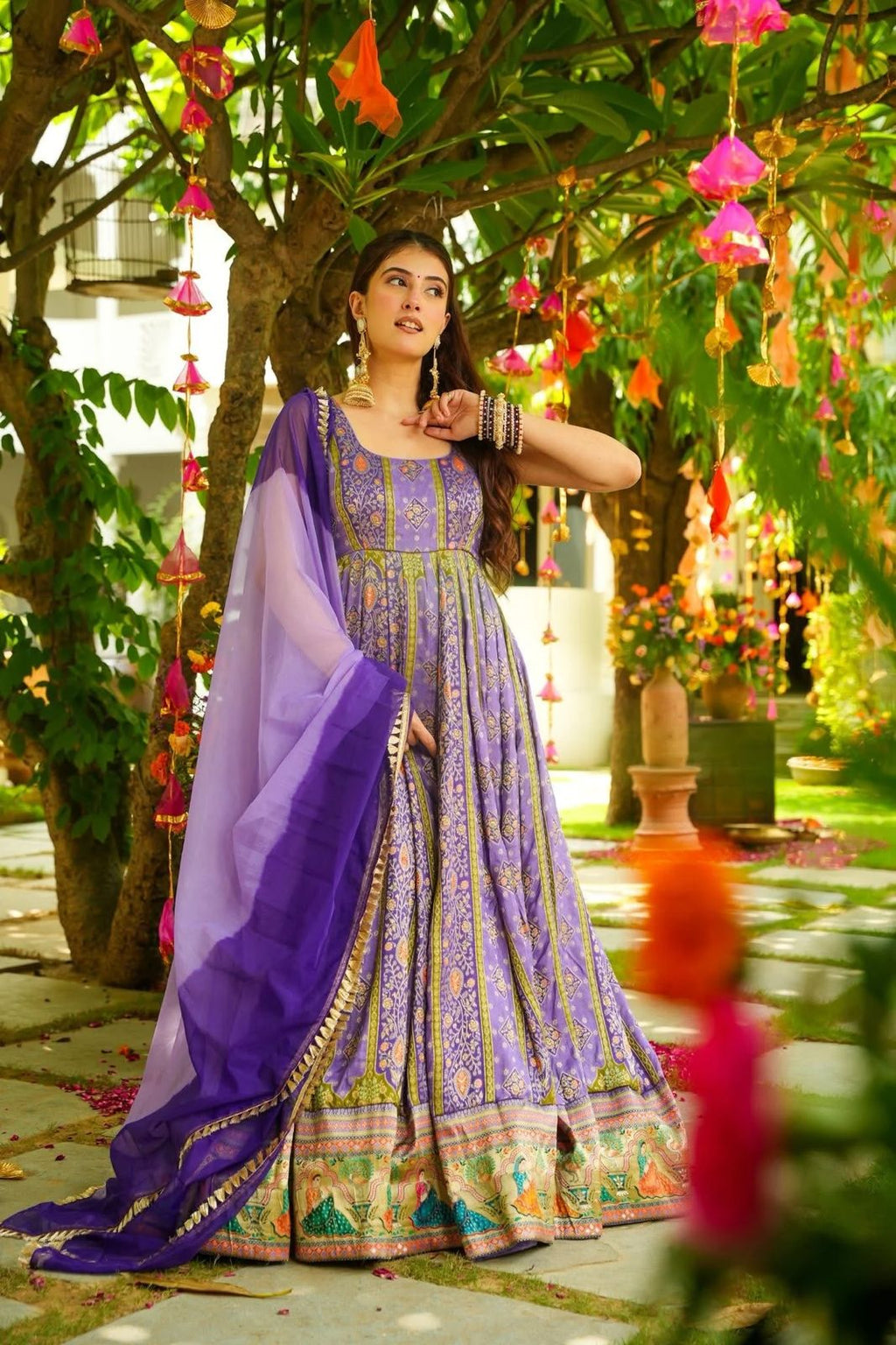 Woman in a purple and multicolored Anarkali Gown standing in a garden with flowers and trees.
