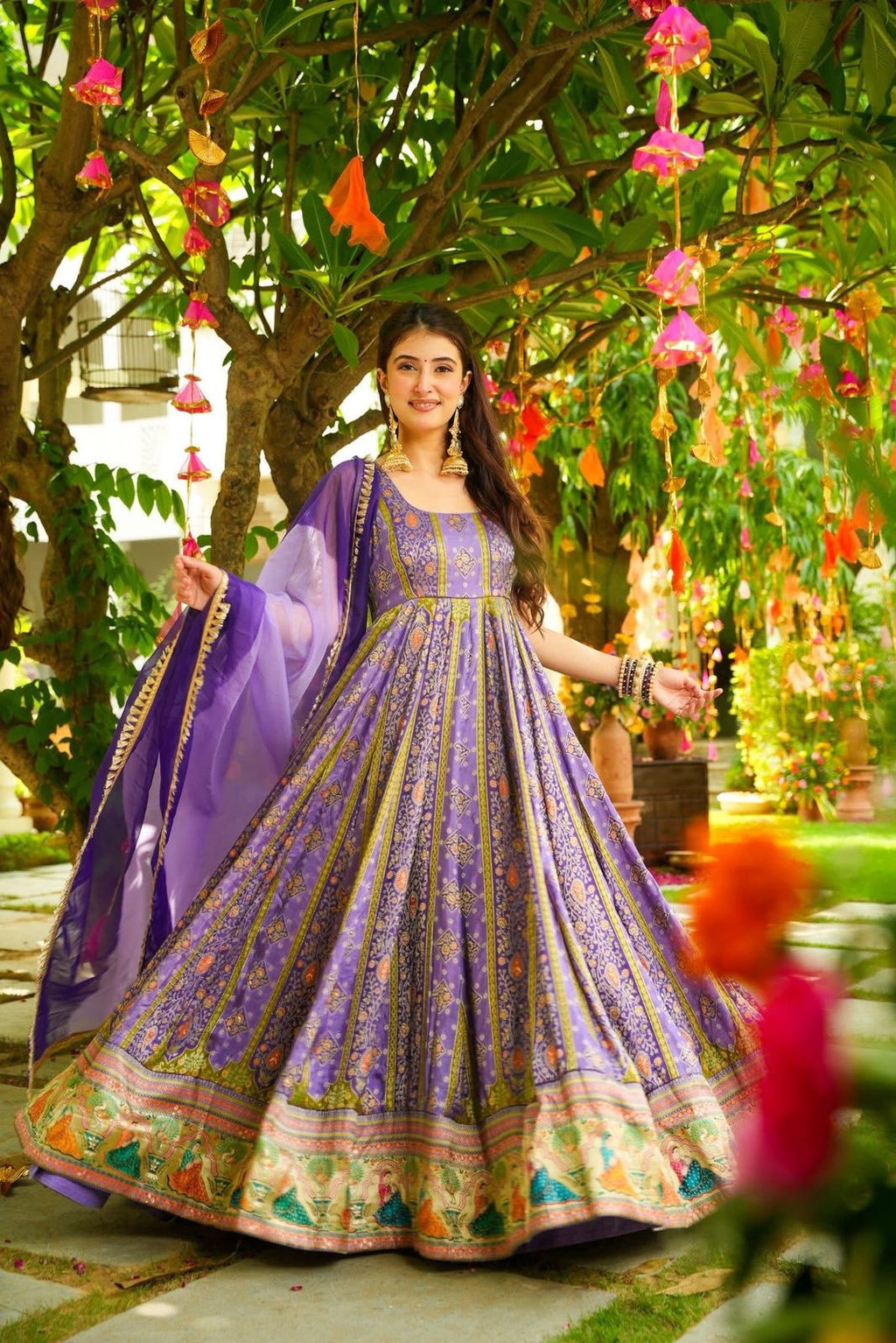 Woman in a purple and green Anarkali Gown standing in a garden with flowers and trees.