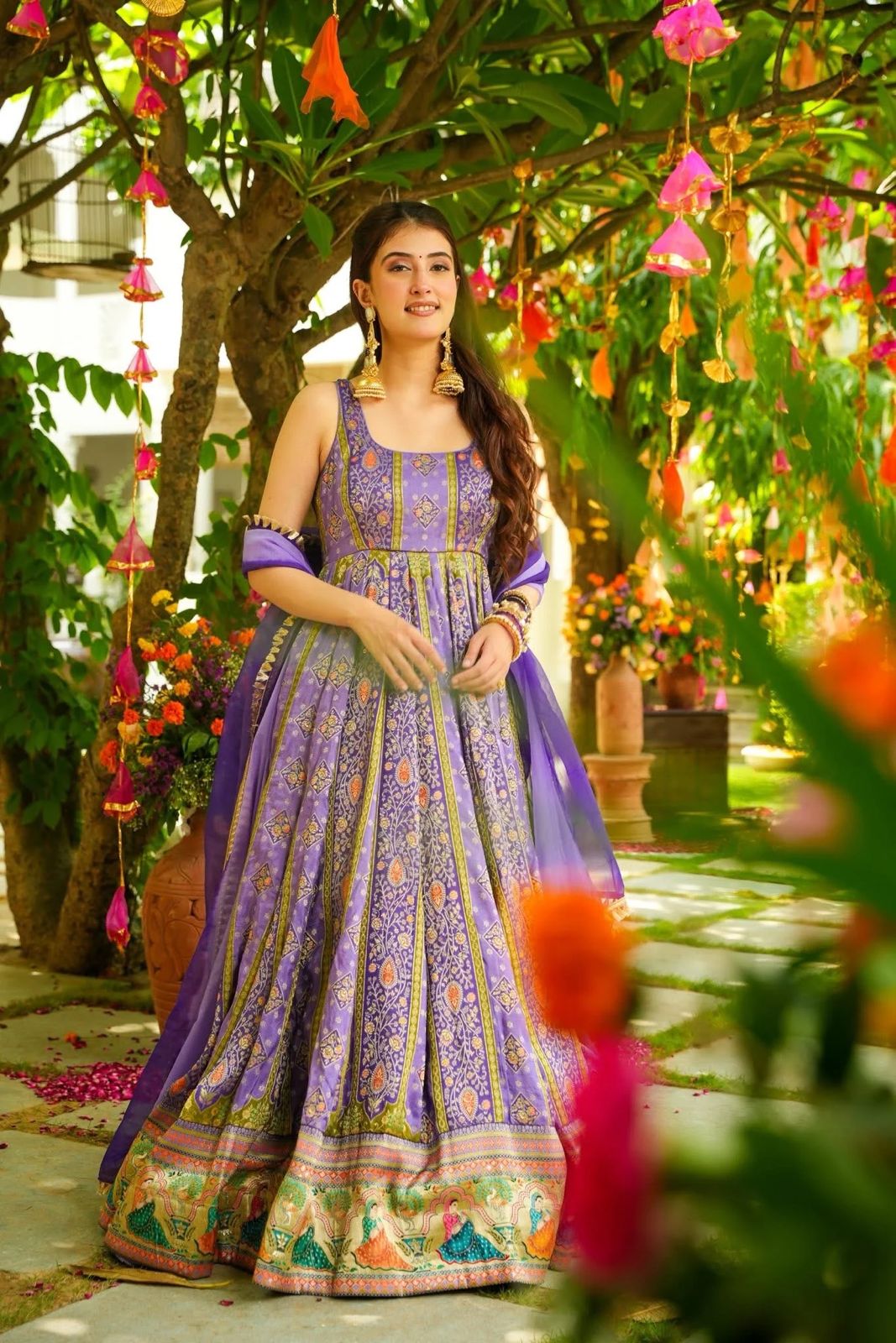 Woman in a purple and green Anarkali Gown outfit standing in a garden with flowers and trees.