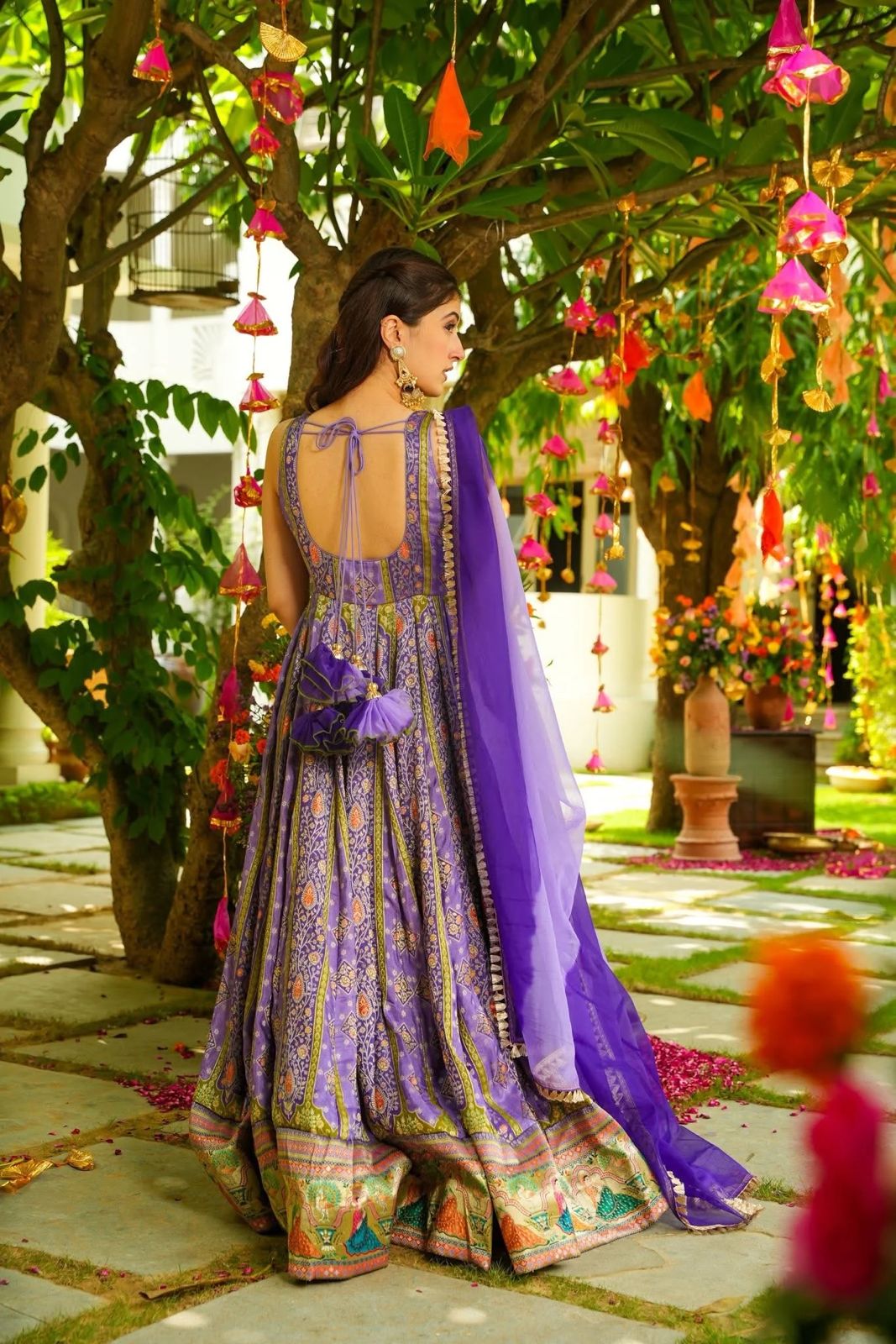 Woman in a Anarkali Gown outfit standing under a decorated tree with colorful lights and flowers.