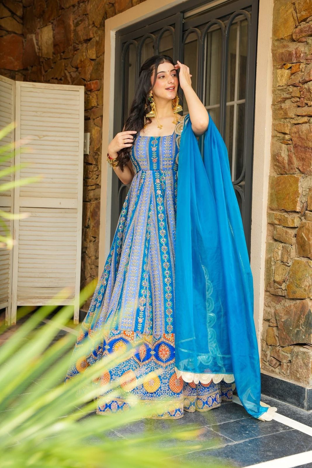 Woman in a blue Anarkali Gown with a blue dupatta standing in front of a stone wall.