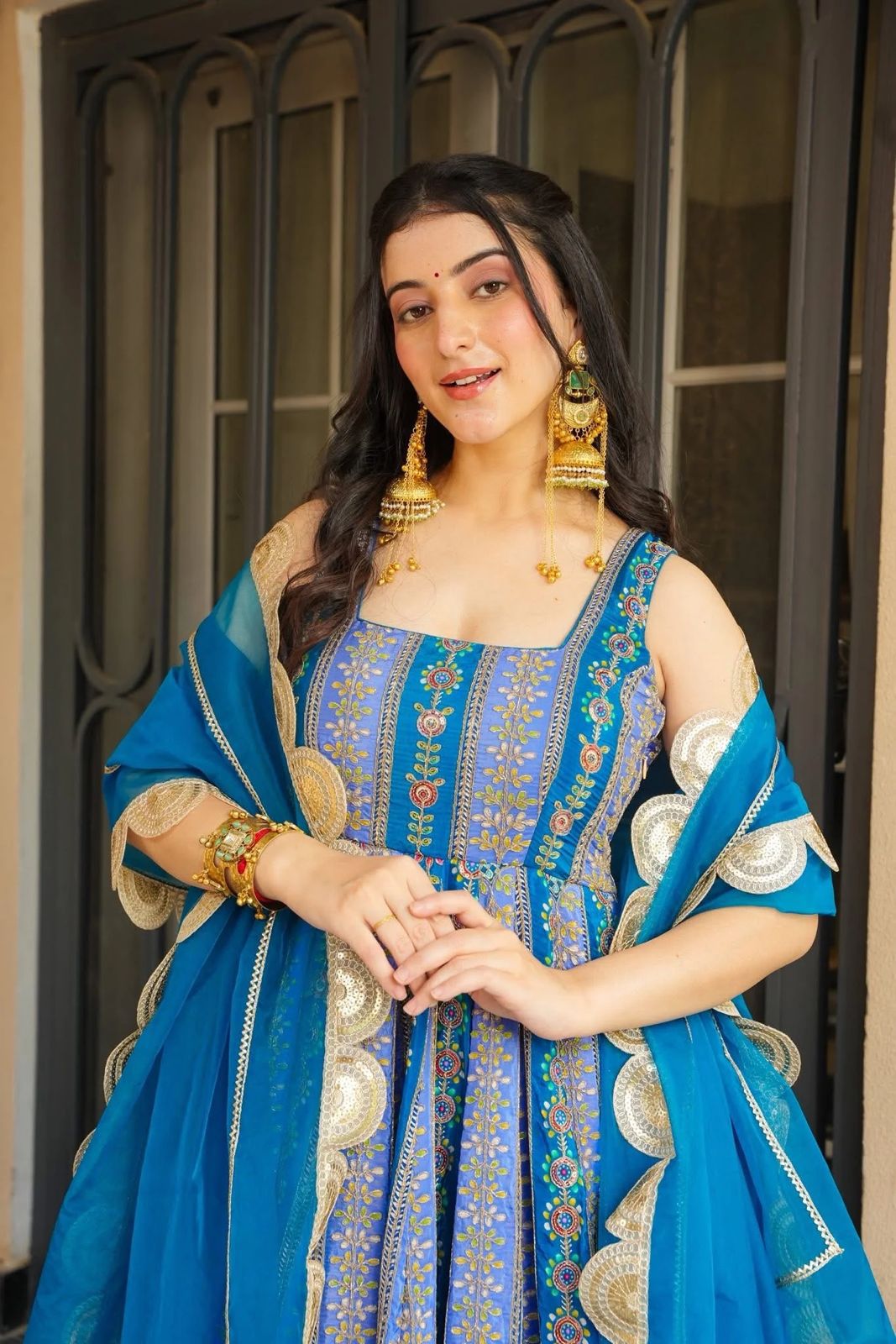 Woman in a blue Anarkali Gown outfit with gold jewelry standing in front of a dark door.