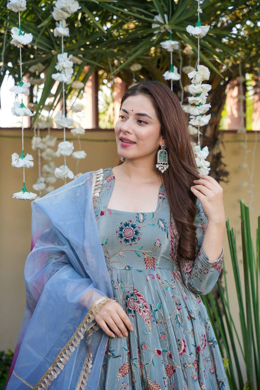 Woman in a Anarkali Gown with a blue dupatta standing in front of hanging decorations.