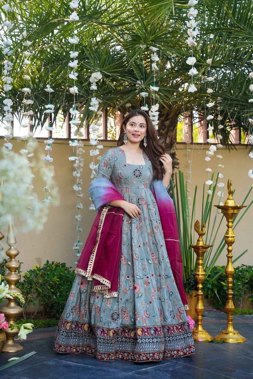 Woman in a Anarkali Gown outfit with floral patterns and a pink dupatta, standing outdoors with greenery and decorative elements.