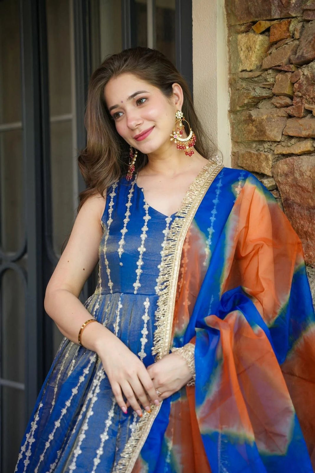 Woman in a blue and orange Anarkali Gown outfit standing against a stone wall.