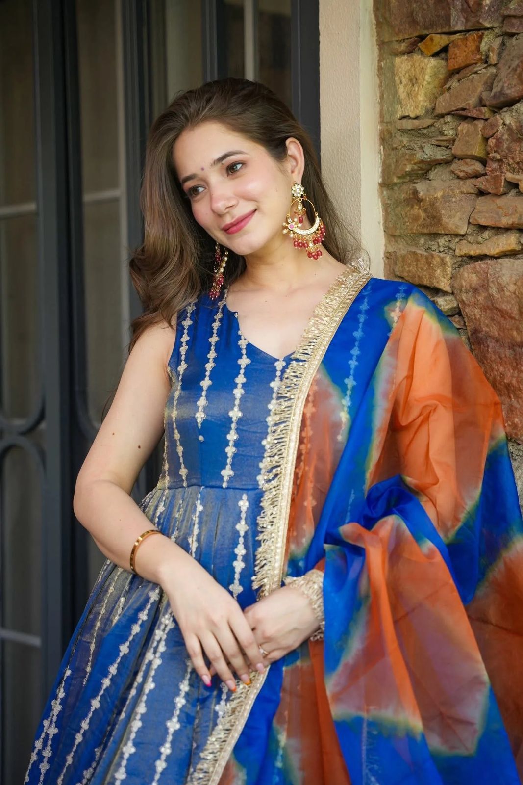 Woman in a blue and orange Anarkali Gown outfit standing against a stone wall.