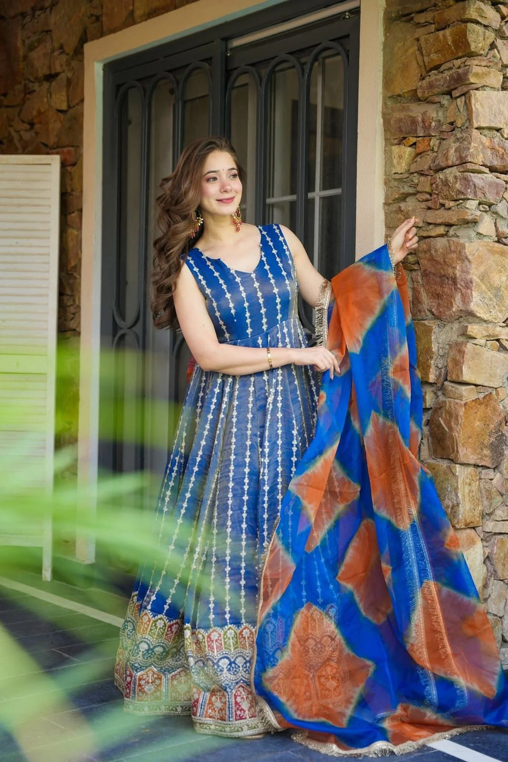 Woman in a blue Anarkali Gown outfit with a patterned dupatta, standing against a stone wall.