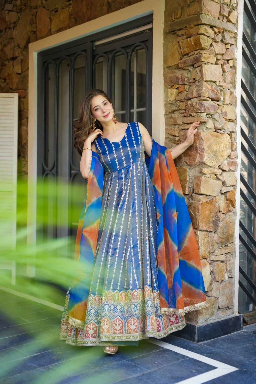 Woman in a colorful Anarkali Gown standing in front of a stone wall.