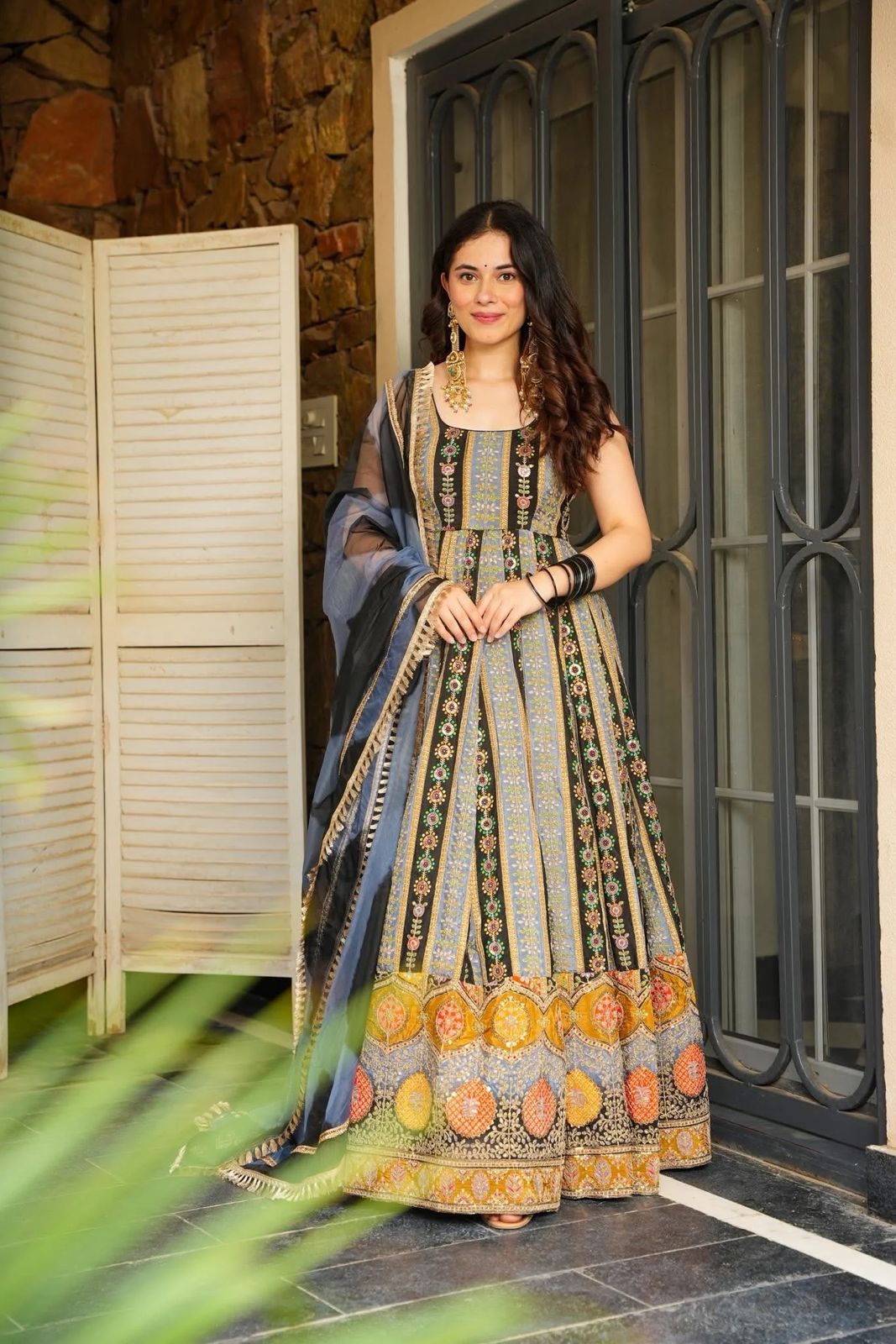 Woman in Anarkali Gown outfit standing in front of a door with stone wall background